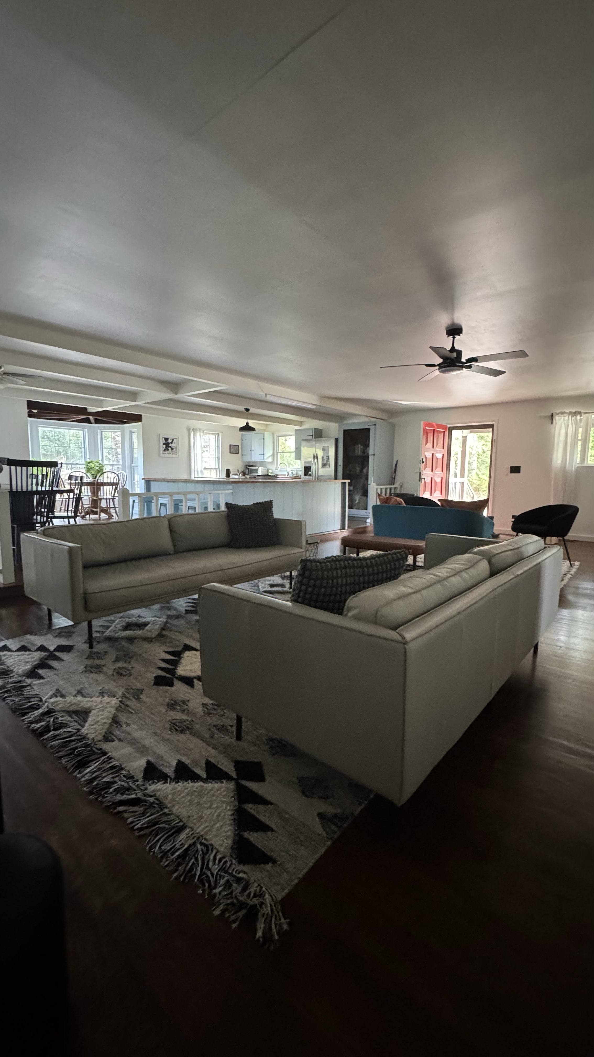 Living room with two beige sofas, a black armchair, a patterned rug, and a blue armchair in the background near large windows and a red front door. The room has wooden flooring and a ceiling fan.