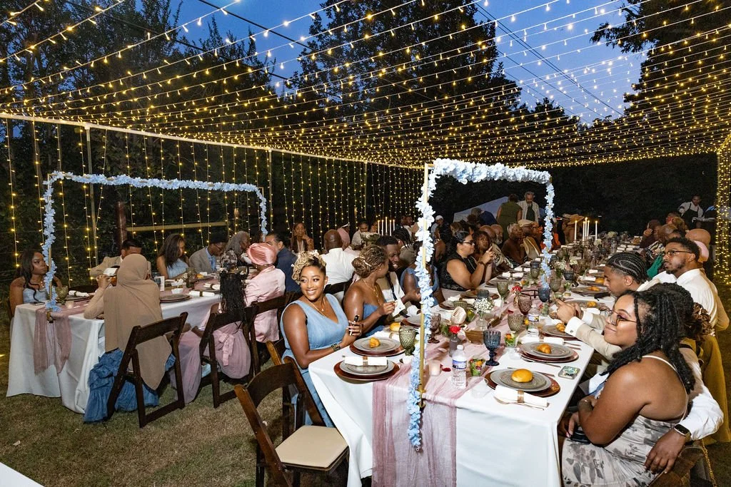 People gathered at a dinner party under string lights with decorated tables and floral accents.