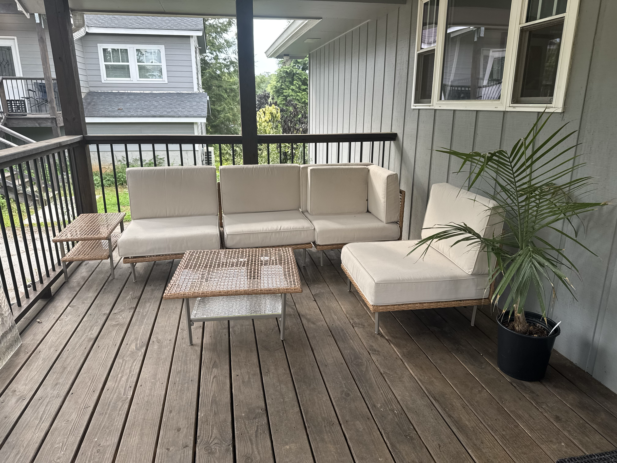 A backyard porch with outdoor furniture including a sectional sofa and a right chaise lounge, a wooden coffee table, a small side table, and a potted palm plant.