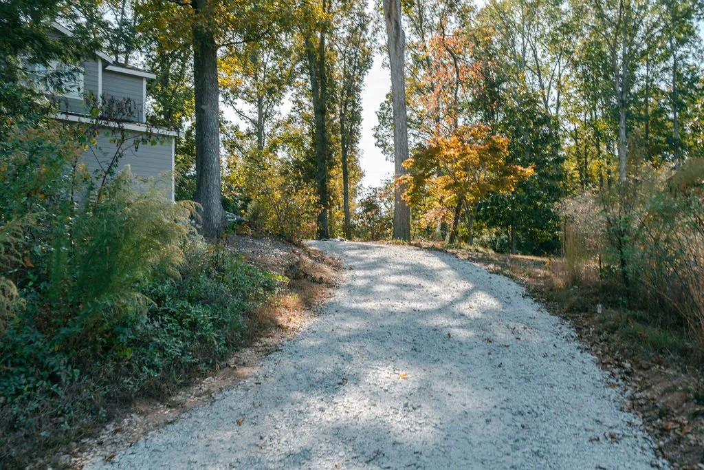 A gravel driveway winding through a wooded area with trees showing fall foliage and a house partially visible on the left.