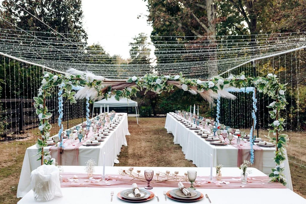 Outdoor wedding or event setup with long tables covered in white tablecloths, decorated with pink runners, candles, and floral centerpieces. Overhead string lights and a floral arch frame the tables, with trees and a grassy area in the background.