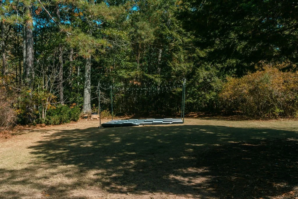 A small outdoor stage with a checkered black and white playing surface, surrounded by trees and bushes, with string lights hanging above.
