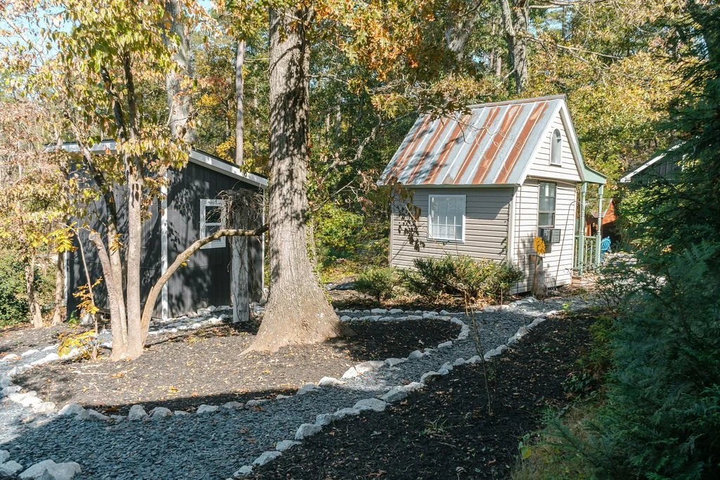 A small house with a rusted metal roof and a porch, surrounded by trees and a garden with a gravel pathway.
