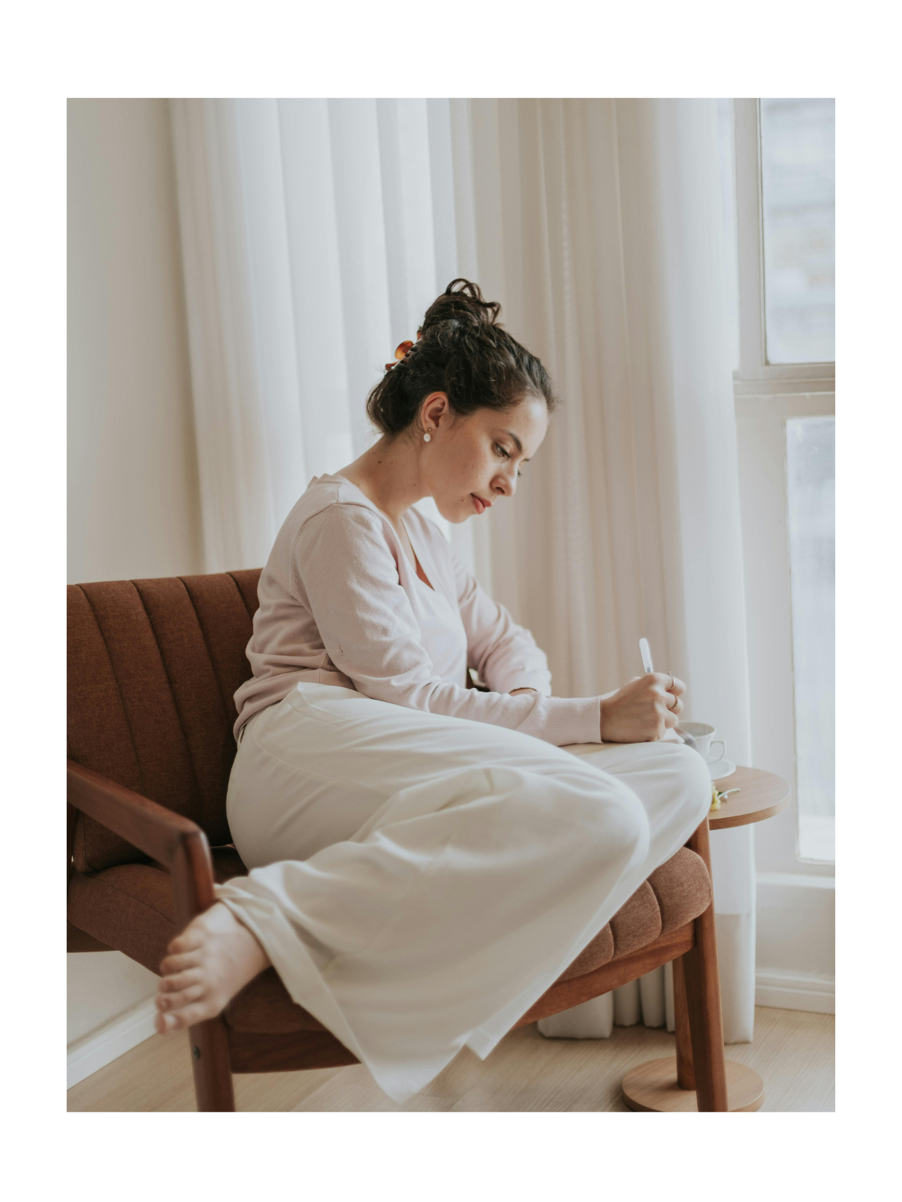 Woman sitting on a brown couch near a window, writing in a notebook.