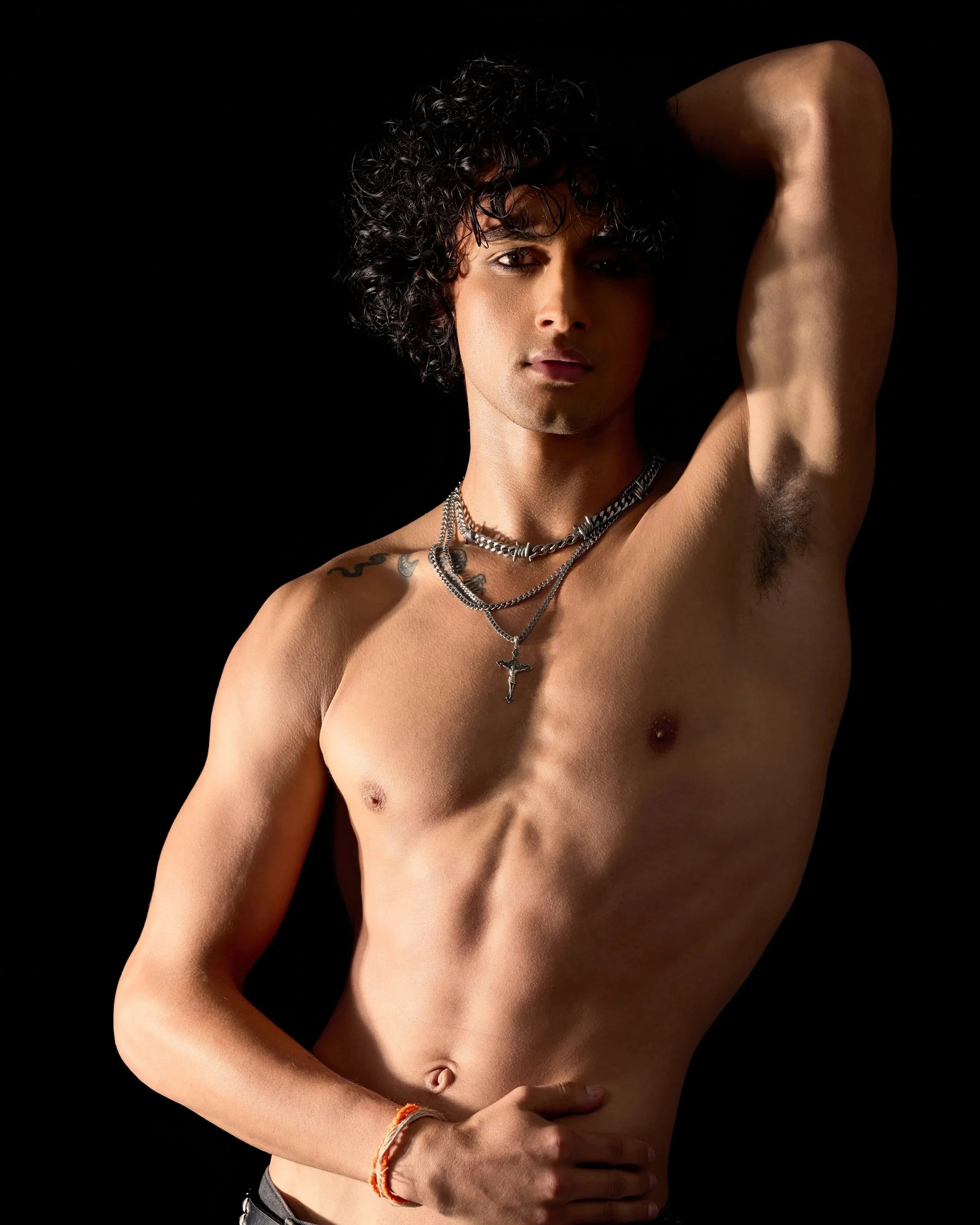 A shirtless young model and actor, Jett Williams, with curly black hair and multiple necklaces, posing with one arm raised and hand behind his head against a black background.