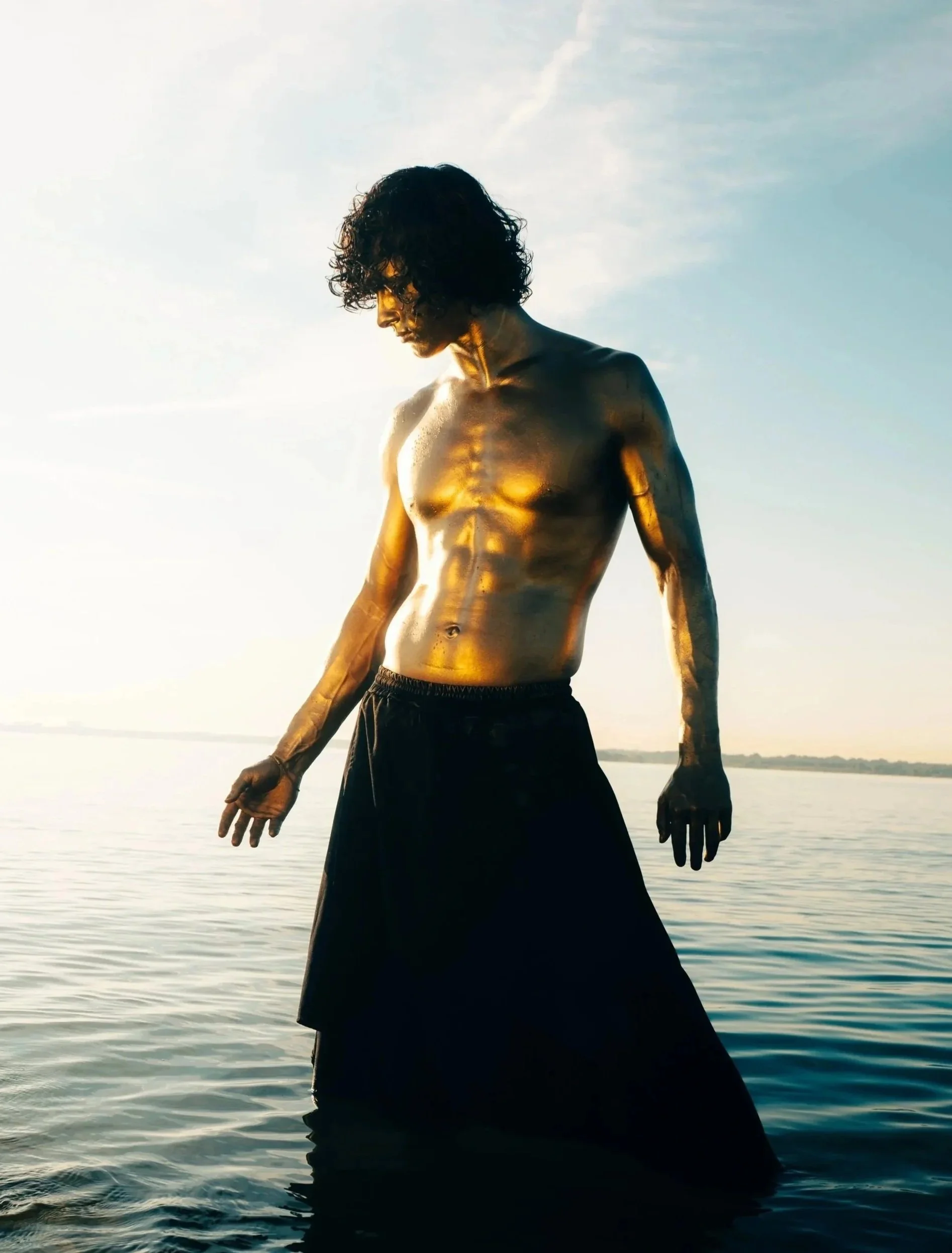 A model and actor, Jett Williams, with curly hair and gold body paint standing in water, wearing black pants, during sunset or sunrise.