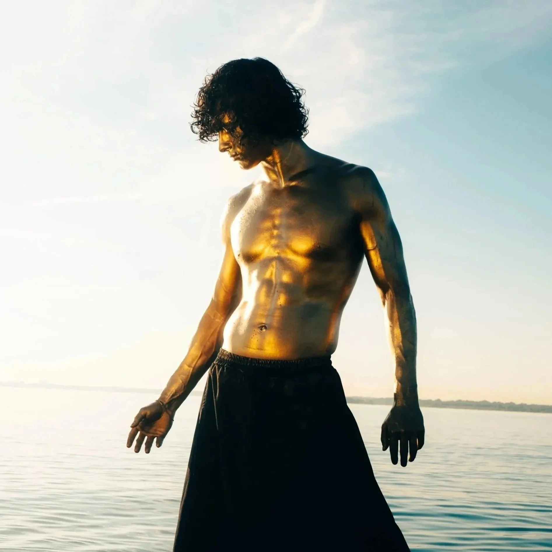 A model and actor, Jett Williams, with curly hair and gold body paint standing in water, wearing black pants, during sunset or sunrise.