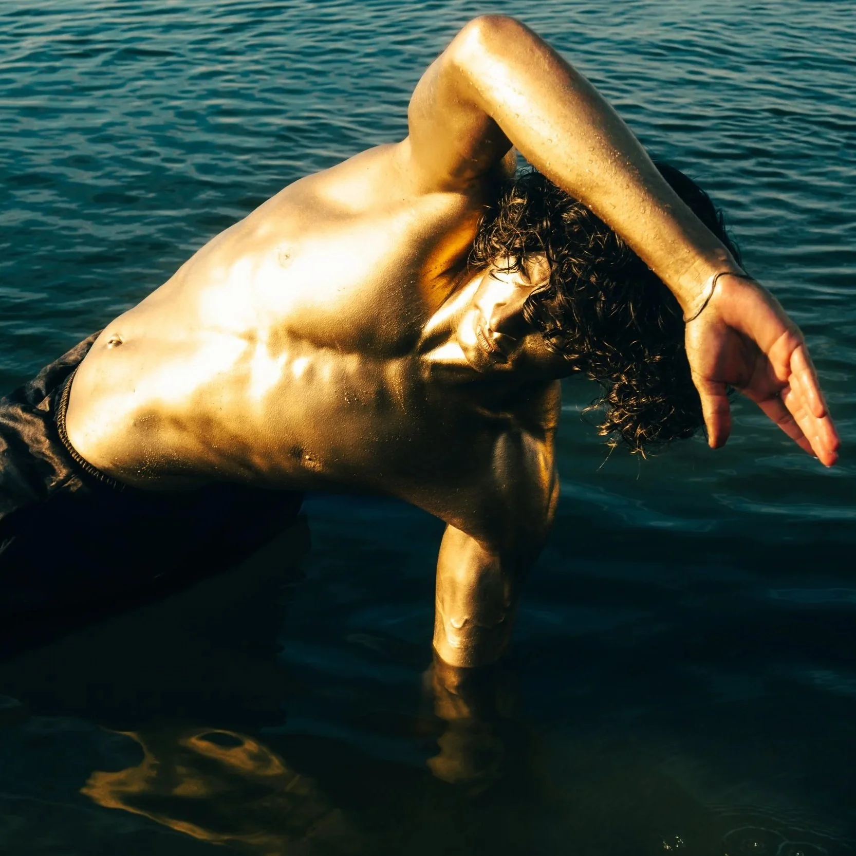 A shirtless model and actor, Jett Williams, with curly hair is modeling in water, covered with gold paint, with one arm bent and head lowered, against a backdrop of water.