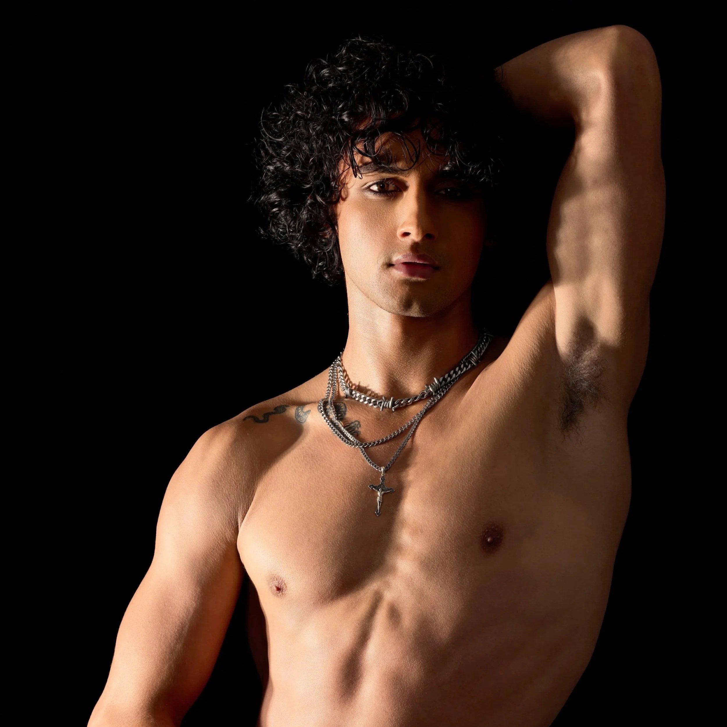 A shirtless young model and actor, Jett Williams, with curly black hair and multiple necklaces, posing with one arm raised and hand behind his head against a black background.
