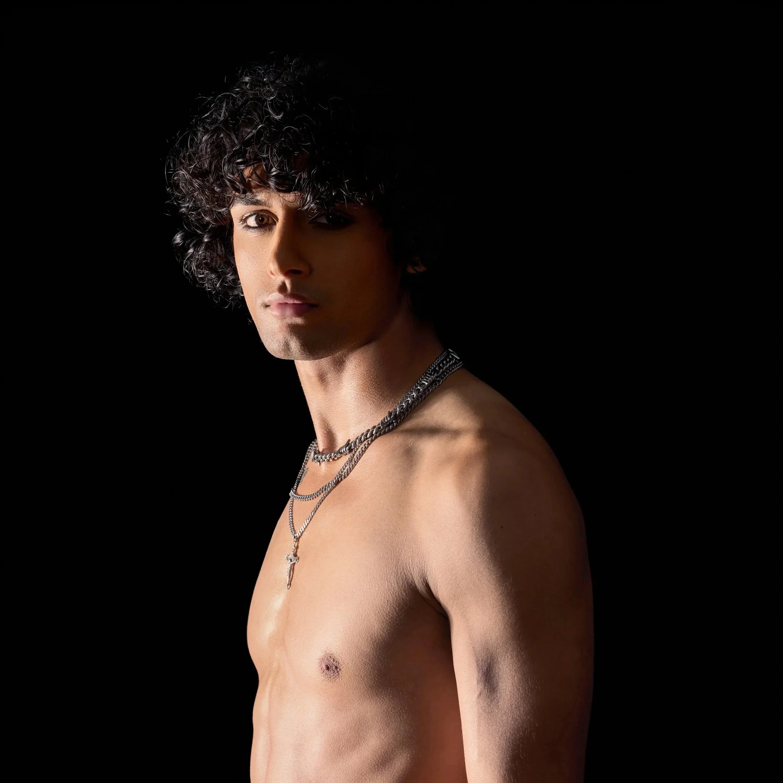 Young model and actor, Jett Williams, with curly dark hair and fair skin, shirtless, wearing silver chains, standing against a black background.