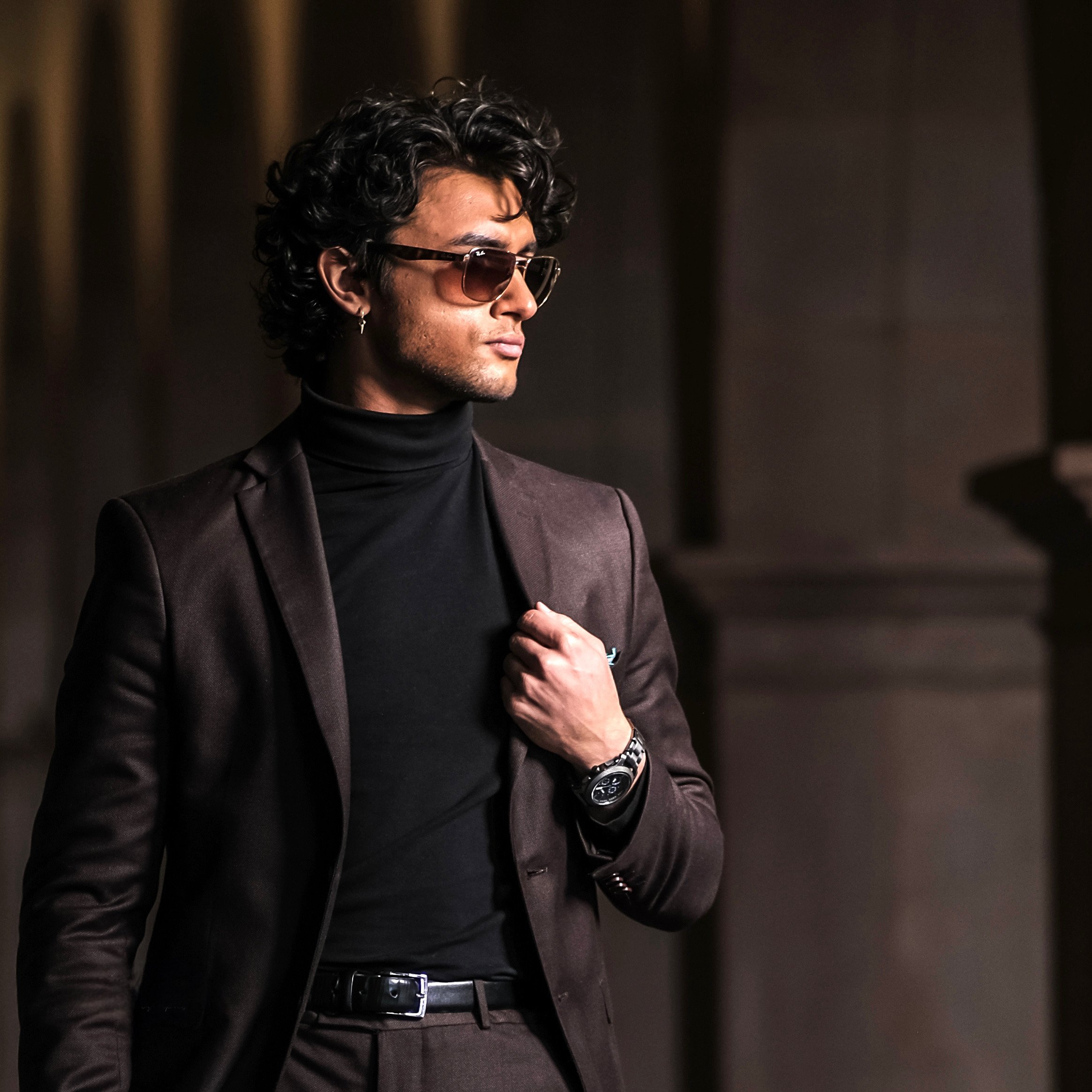 Stylish model and actor, Jett Williams, wearing sunglasses, black turtleneck, and brown blazer, posing indoors with a confident look.