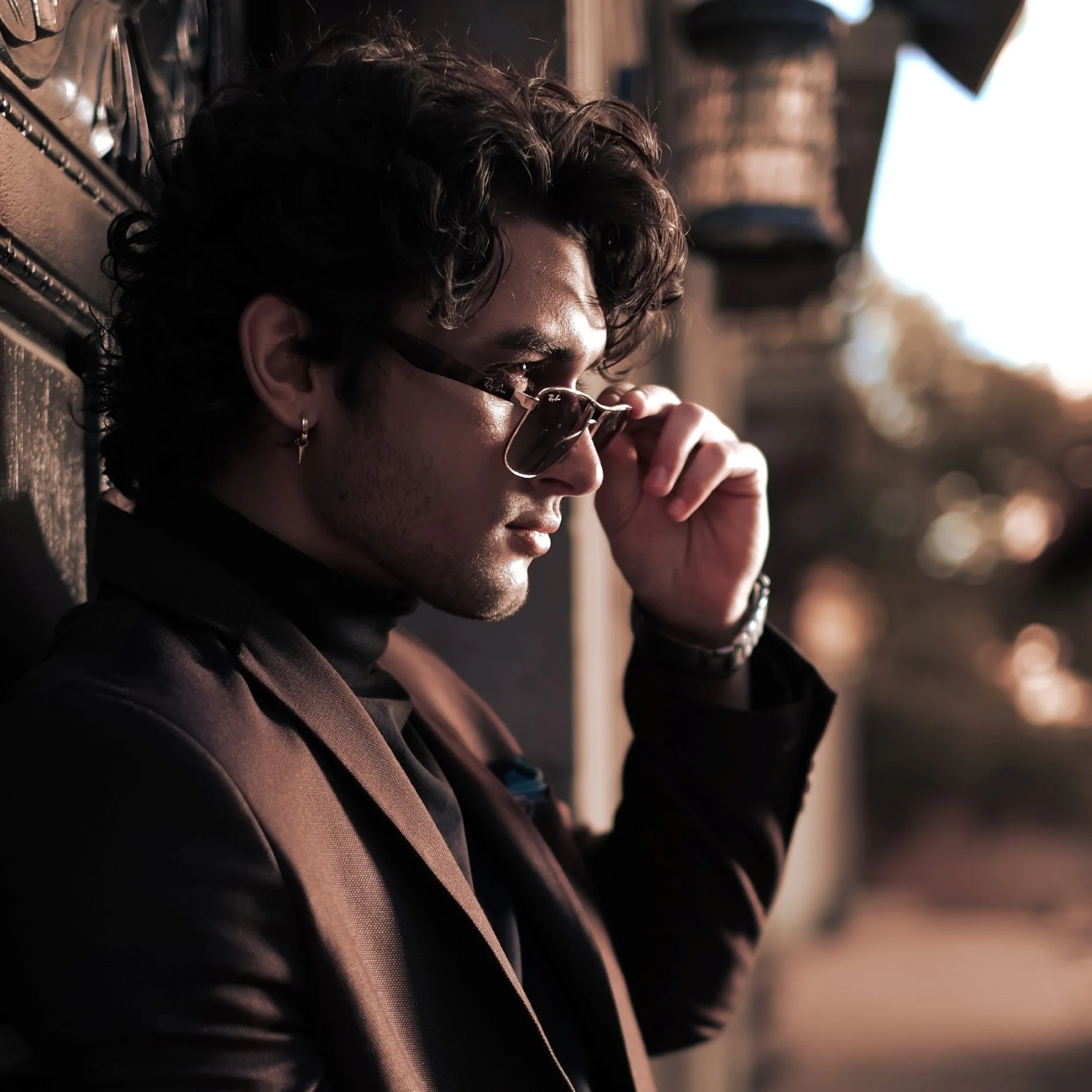 A young model and actor, Jett Williams, with curly dark hair wearing sunglasses, a black turtleneck, and a black blazer, leaning against a wall outdoors during sunset, adjusting his glasses.