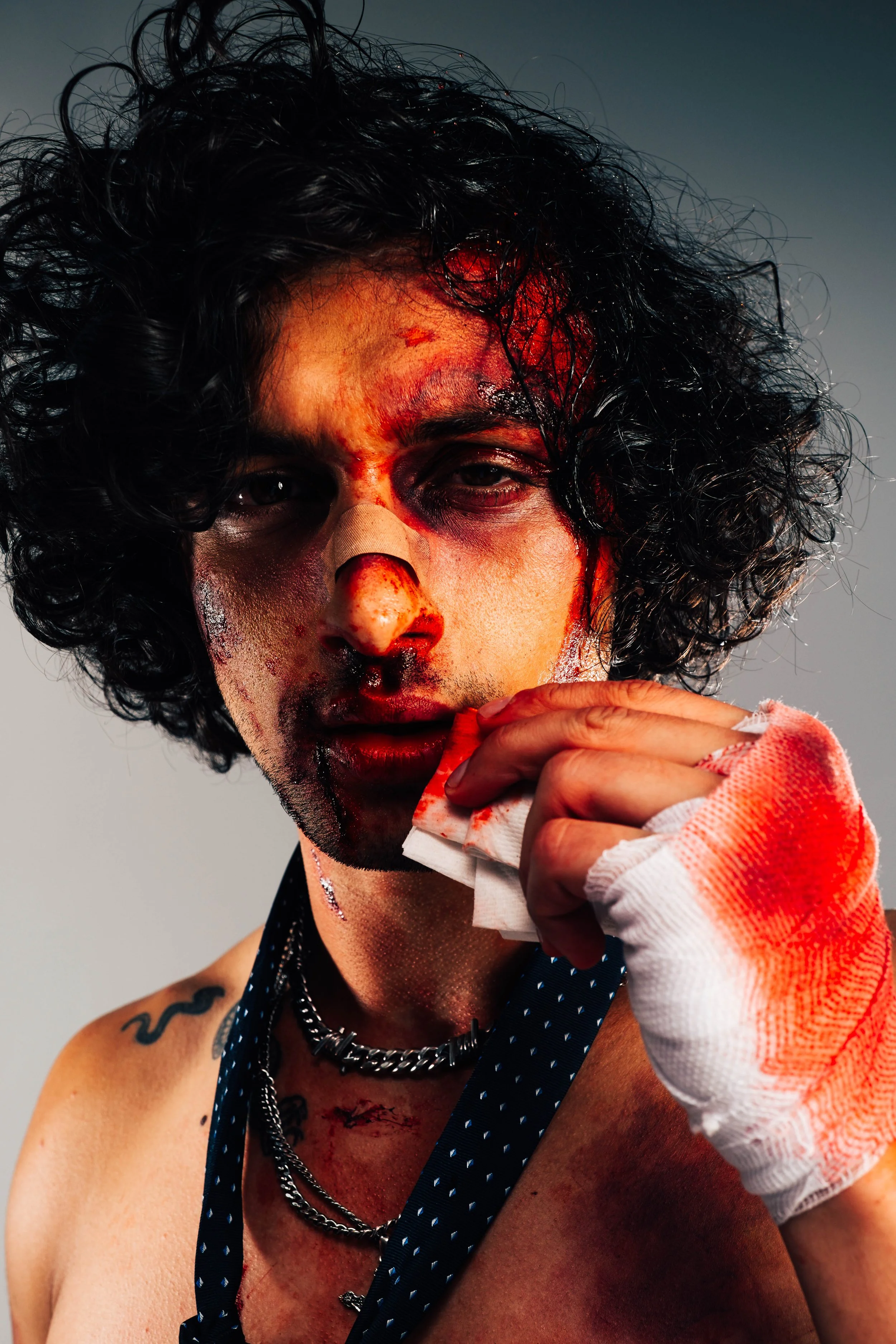 A young model and actor, Jett Williams, with curly hair, blood and injuries on face, holding a bloodied tissue, wearing a sleeveless top and layered necklaces