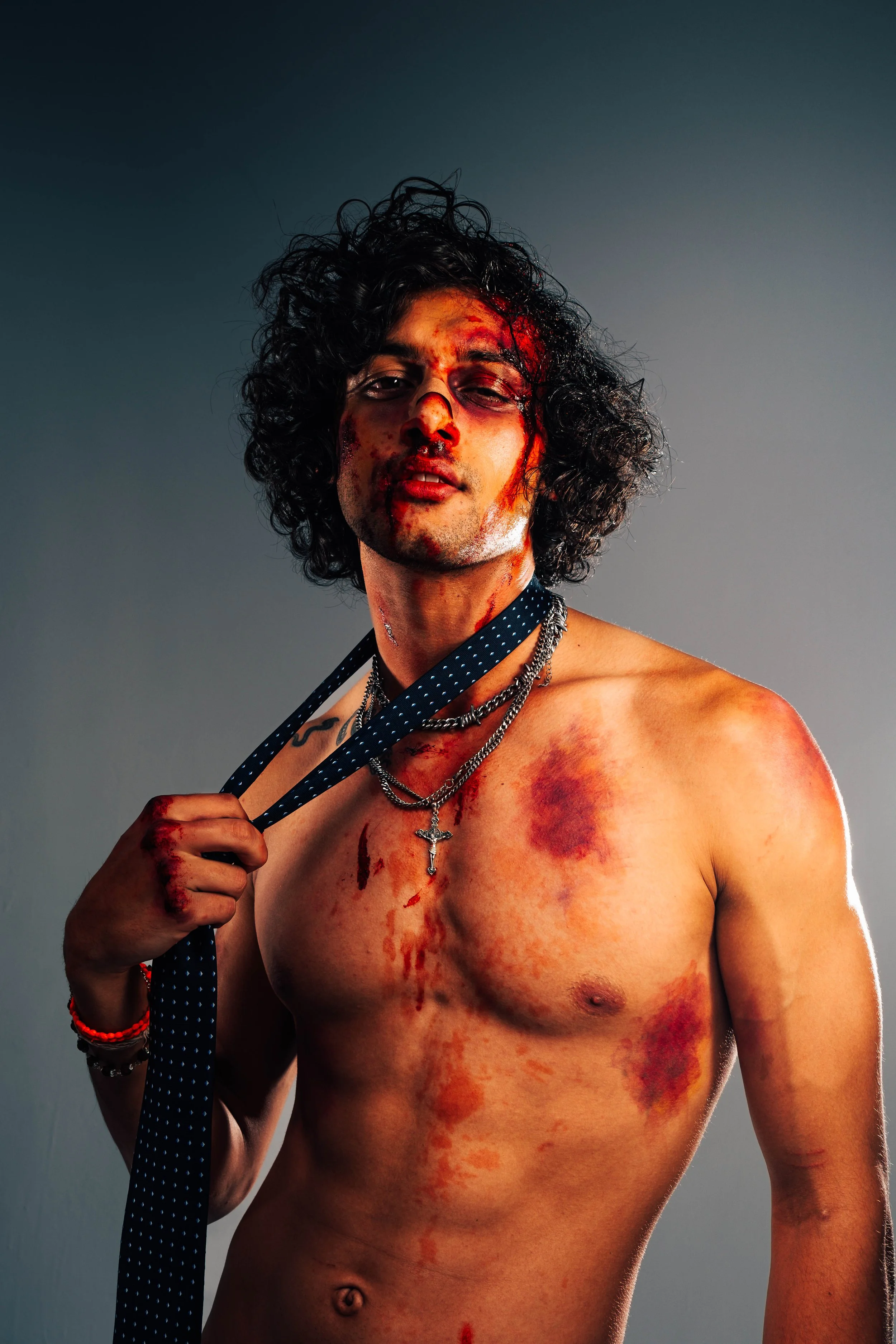 A shirtless young model and actor, Jett Williams, with curly hair, bloodstains, and injuries on his face and body, holding a black tie over his shoulder, against a gray background.