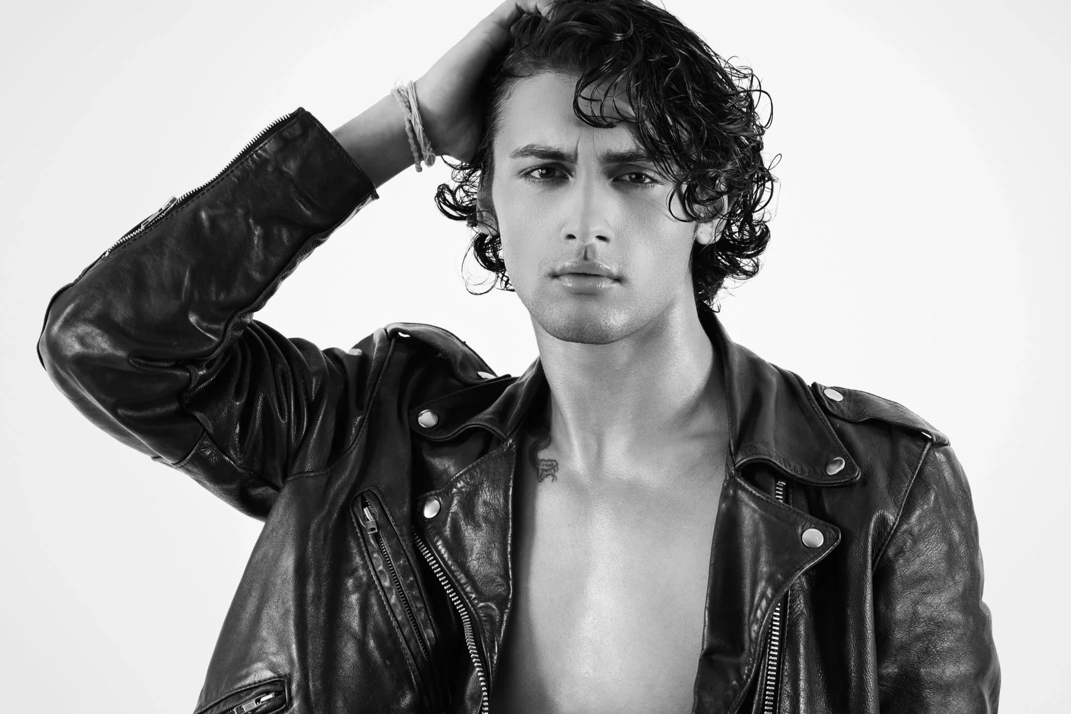 Black and white portrait of a young man with curly hair wearing a leather jacket, looking into the camera with a serious expression, and resting his hand on his head.
