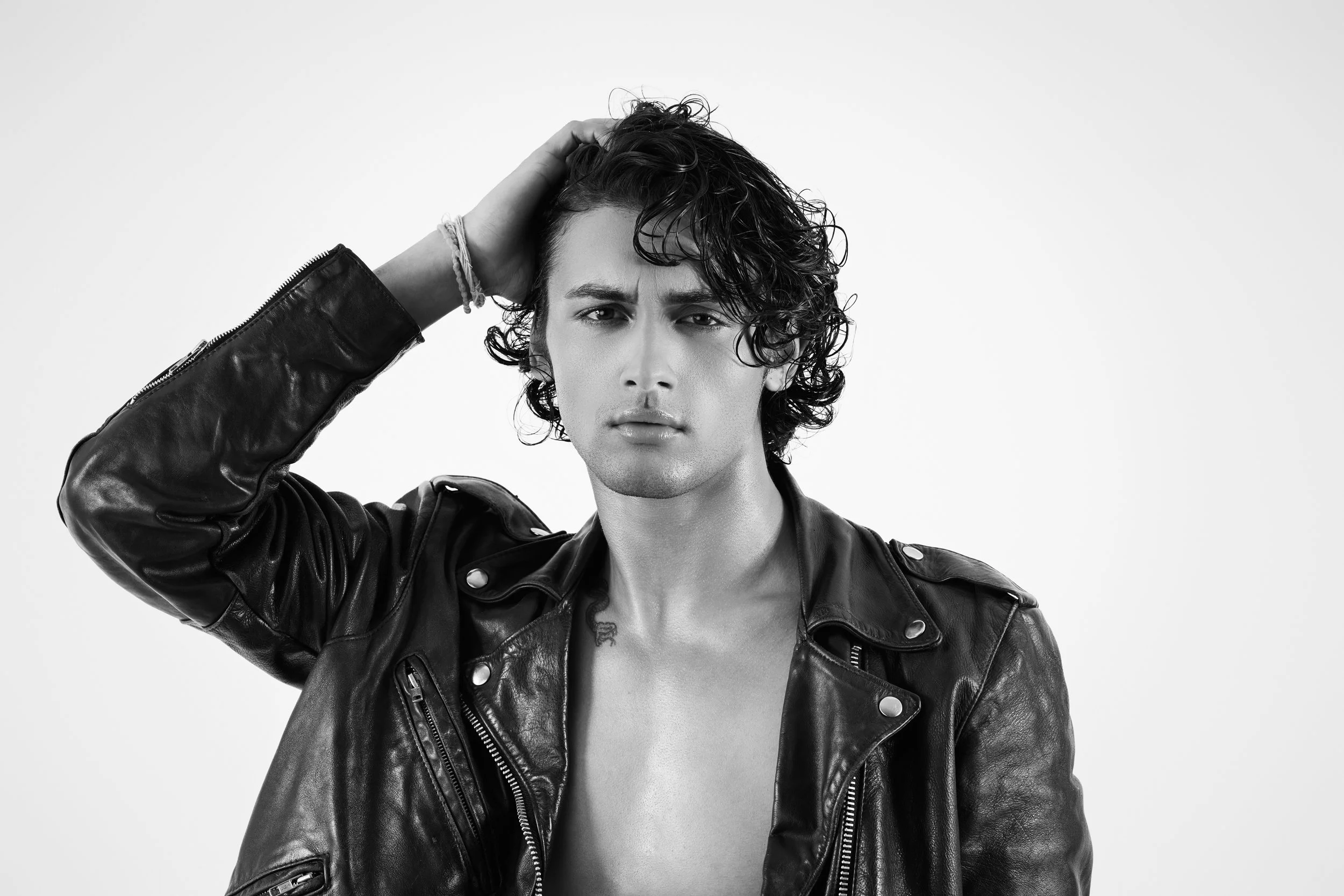 Black and white portrait of young model and actor, Jett Williams, with curly hair, wearing a leather jacket, looking at the camera with a serious expression, hand resting on his head.