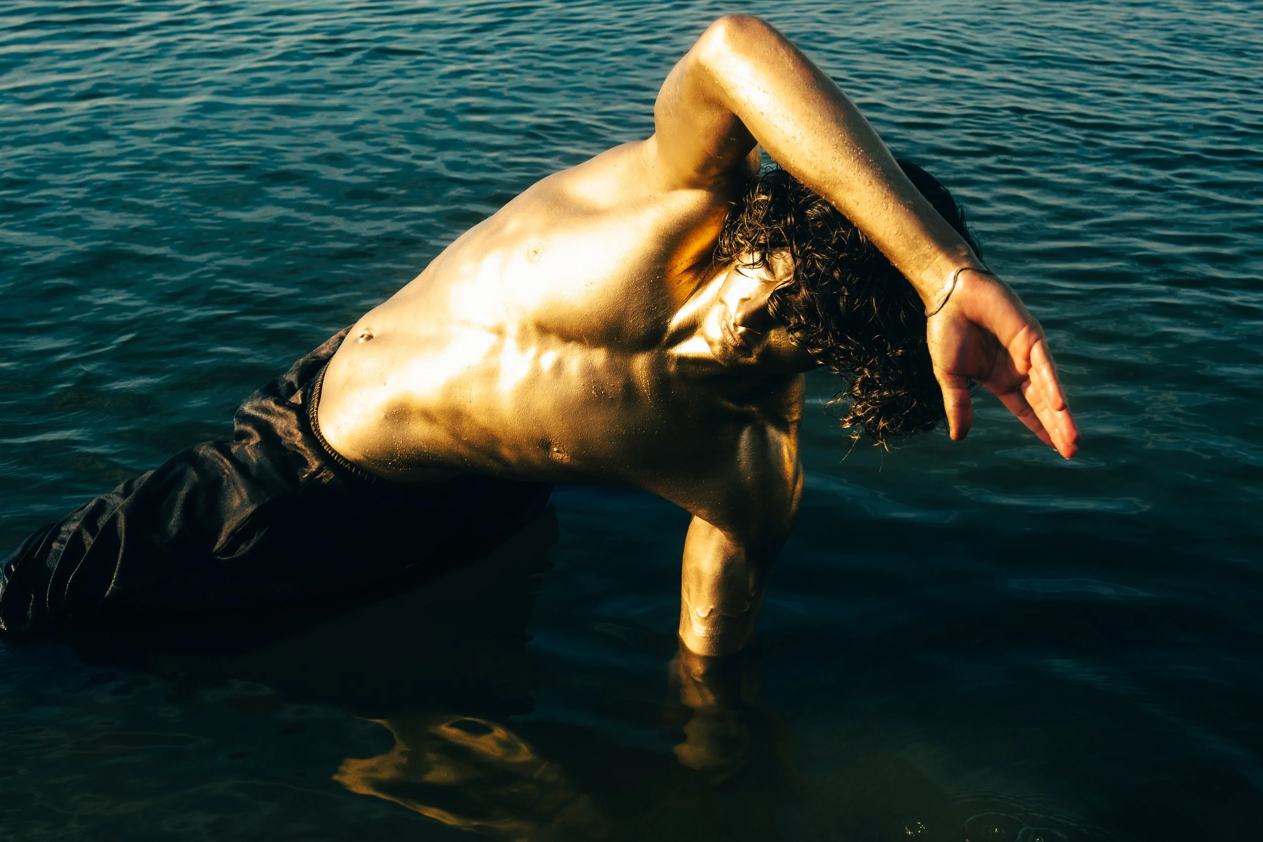 A shirtless model and actor, Jett Williams, with curly hair is modeling in water, covered with gold paint, with one arm bent and head lowered, against a backdrop of water.