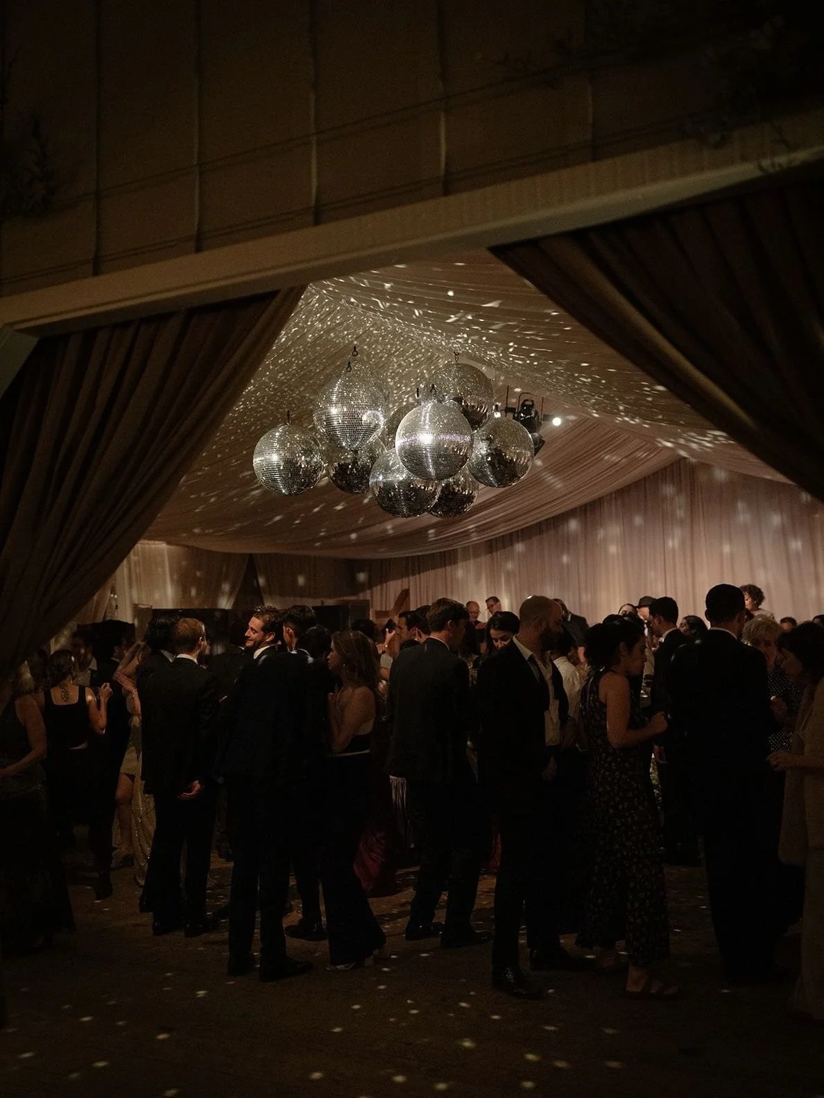 People dancing and socializing at a party with disco balls hanging from the ceiling, creating light reflections across the room.