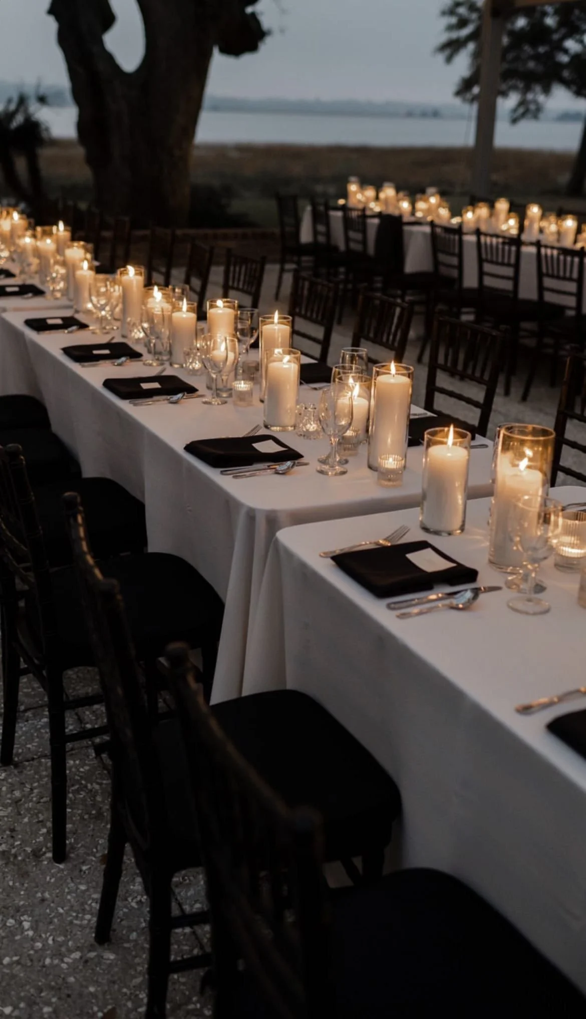 Long banquet table outside at dusk decorated with candles and glassware, overlooking a body of water in the background.