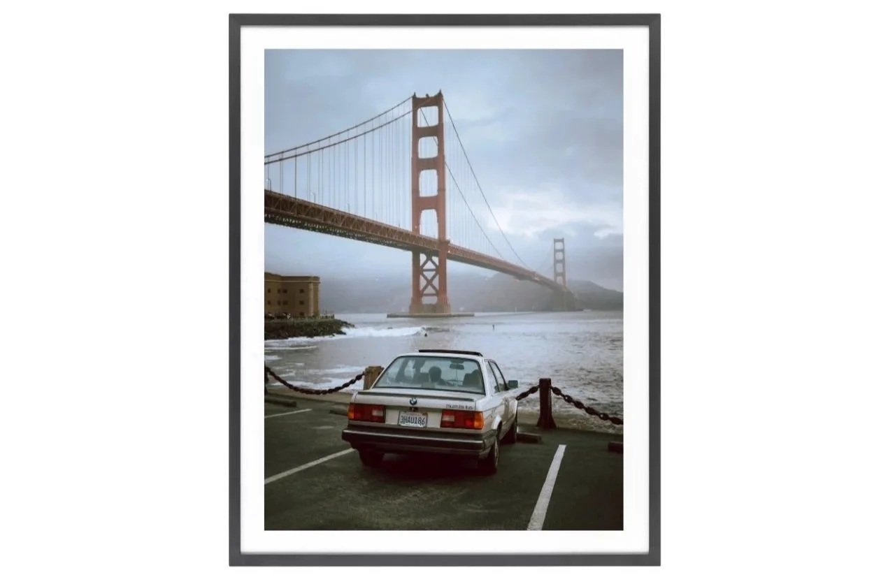Car parked on the road to Fort Point with the Golden Gate Bridge in the background over water on a gloomy, overcast day.