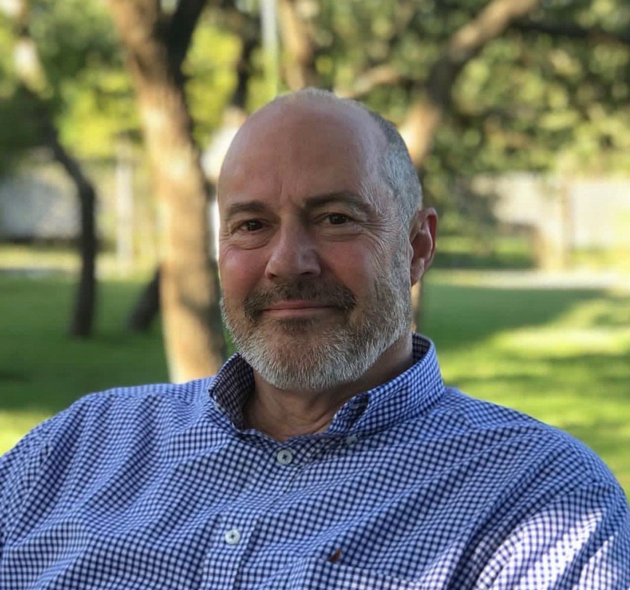 A middle-aged man with a beard and mustache smiling outdoors in a park with trees and green grass in the background, wearing a blue checkered shirt.