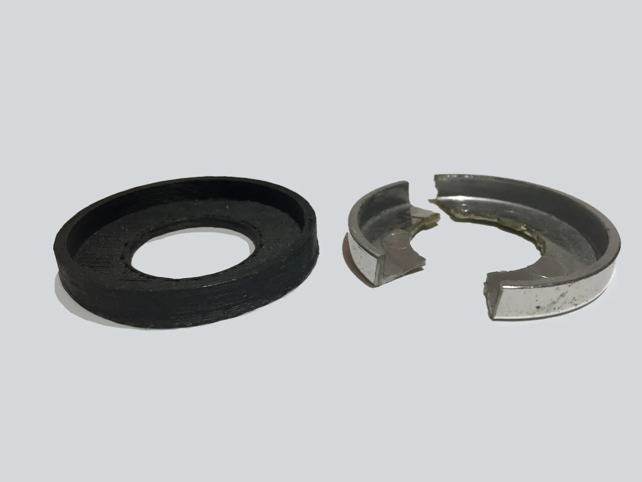 Disassembled ball bearing with a black rubber seal, a metallic cage, and a metallic ring on a plain light gray background.