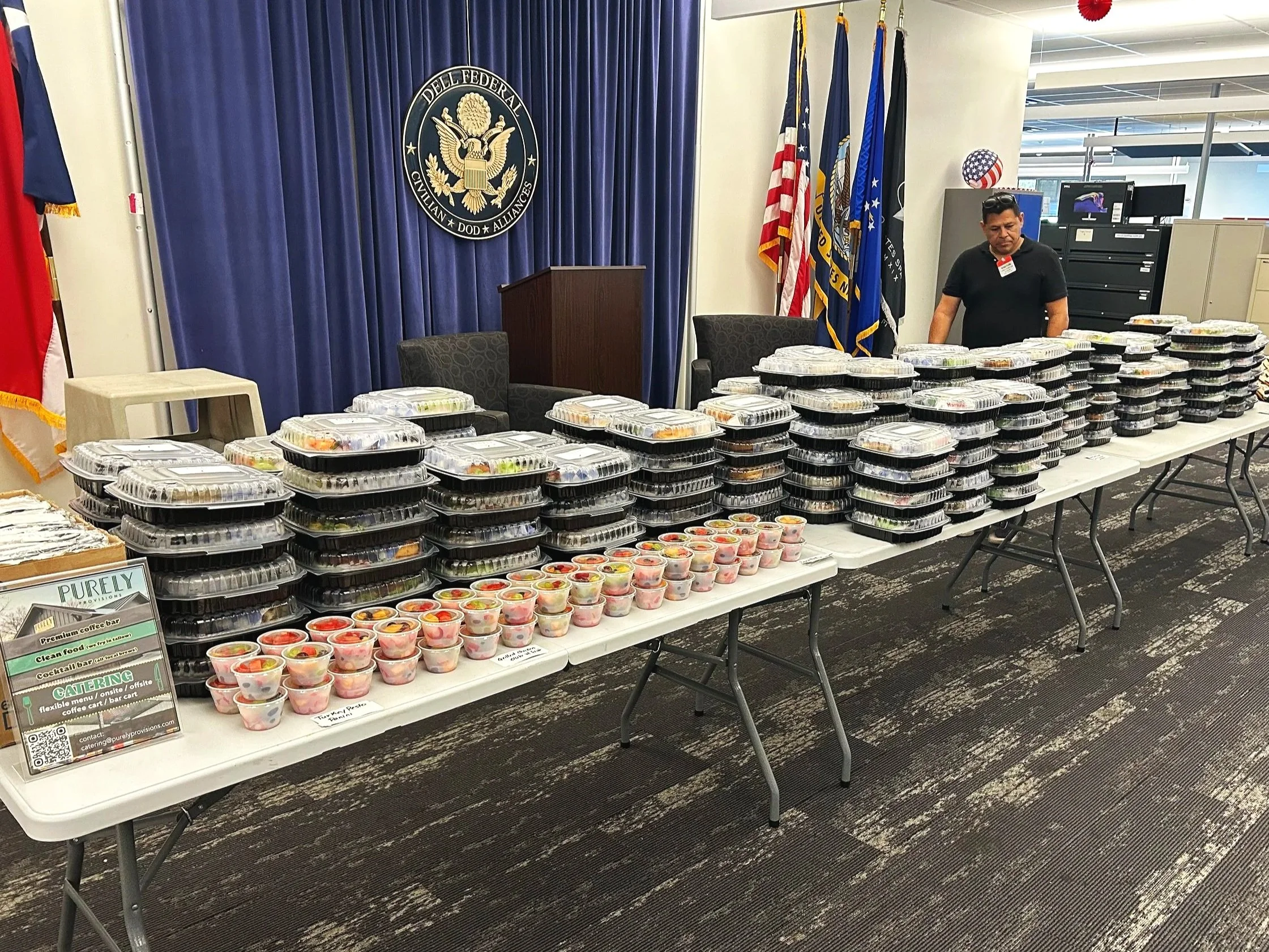 Table with multiple stacked black plastic food containers with clear lids, filled with salads and meals, set up in front of a blue curtain with a government seal and flags, in a conference or meeting room.