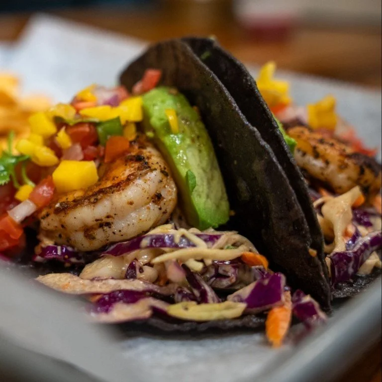 Two tacos filled with grilled shrimp, sliced avocado, chopped tomatoes, and shredded purple cabbage.