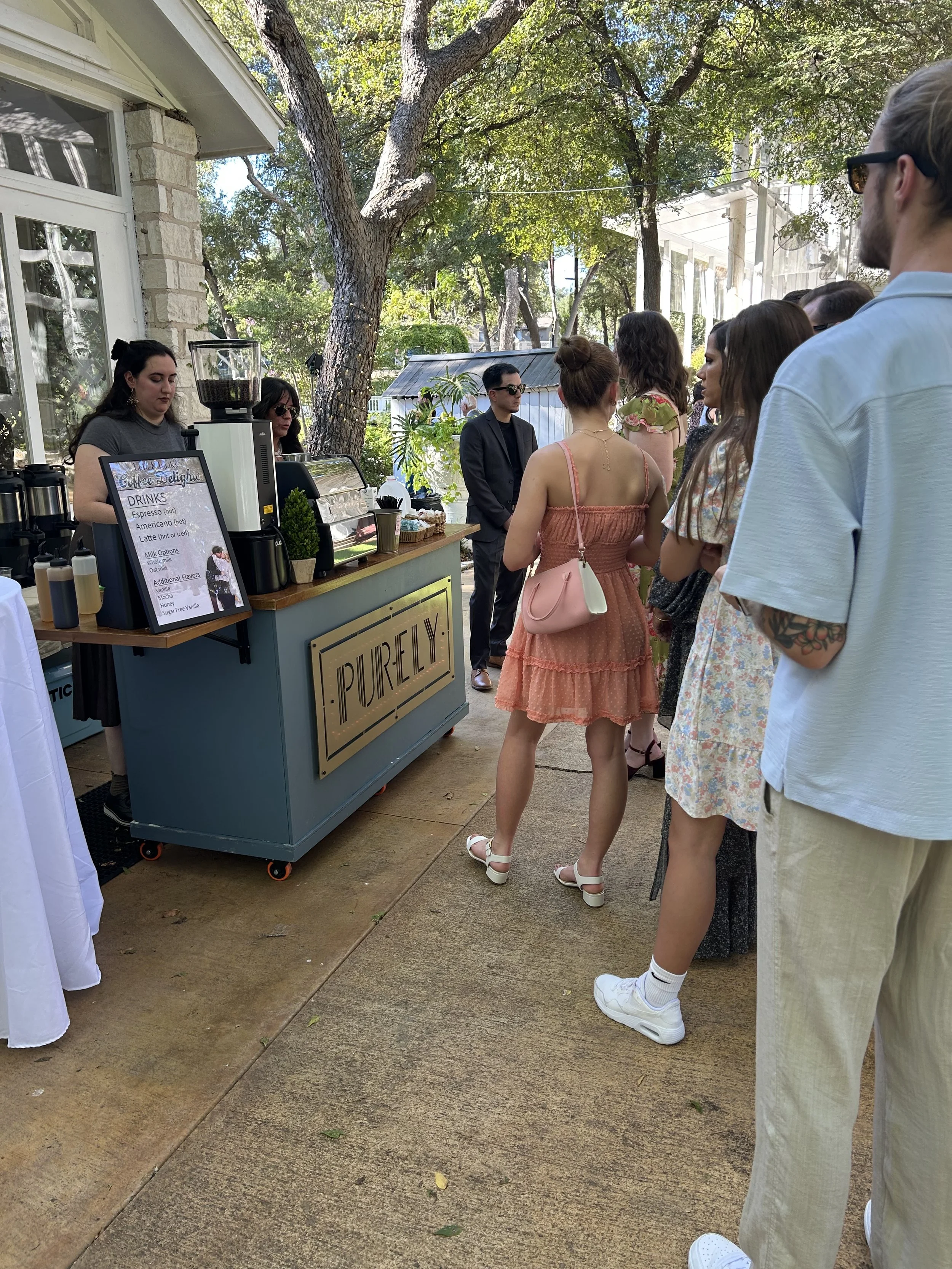 Line of people waiting at an outdoor coffee stand with a barista making drinks, surrounded by trees and sunlight.
