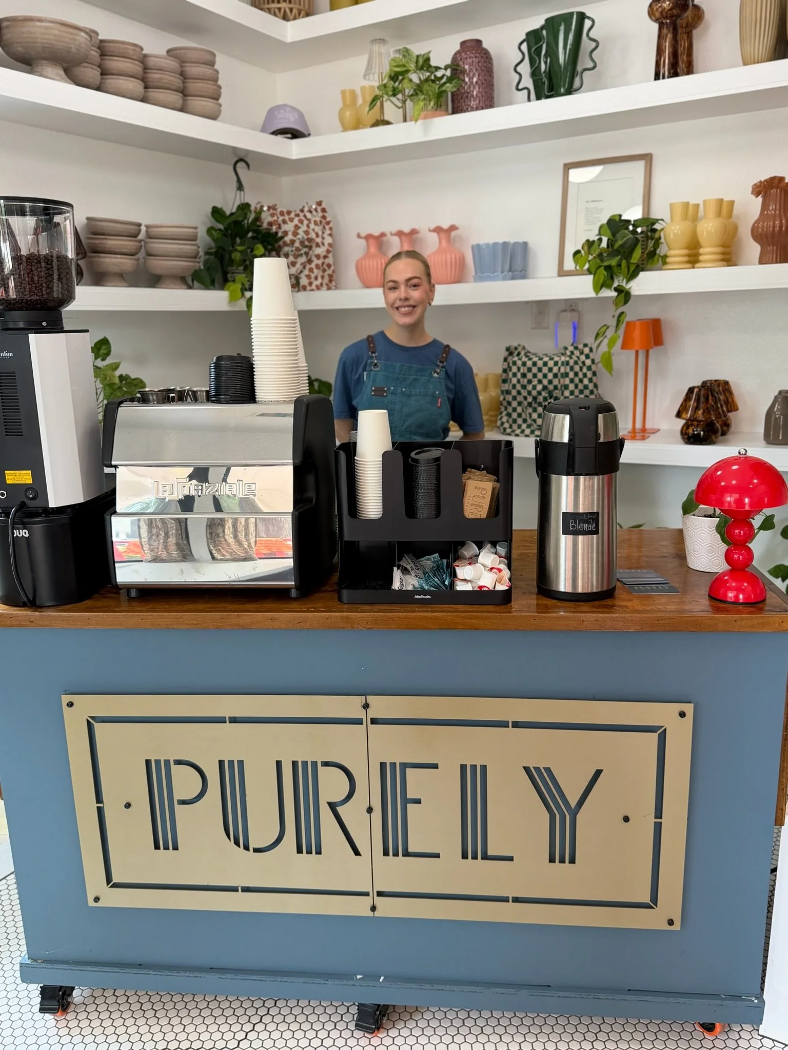 Counter with coffee equipment and a woman smiling behind it, with shelves of decorative pottery and plants in the background.