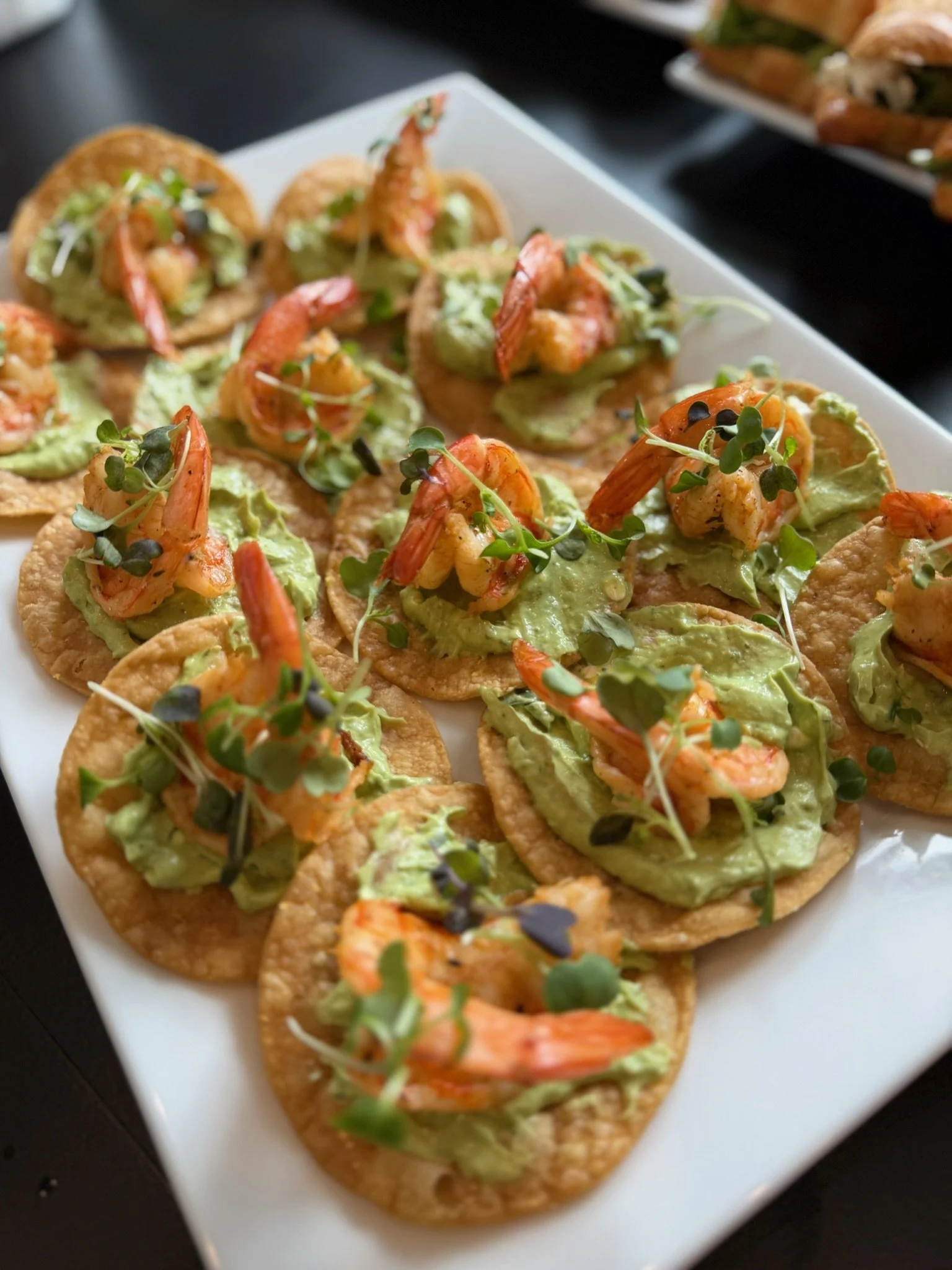 Small round crackers topped with shrimp, avocado, microgreens, and a creamy green sauce on a white rectangular serving platter.