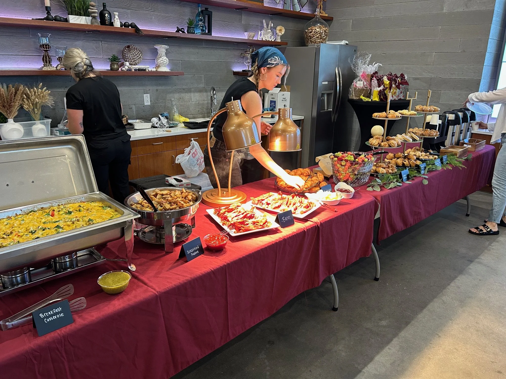 A buffet table with various breakfast dishes such as casserole, roasted potatoes, fruit, and pastries, set on a red tablecloth. There are people serving themselves, and the setting appears to be in a cozy restaurant or cafe with decorated shelves and a silver refrigerator in the background.