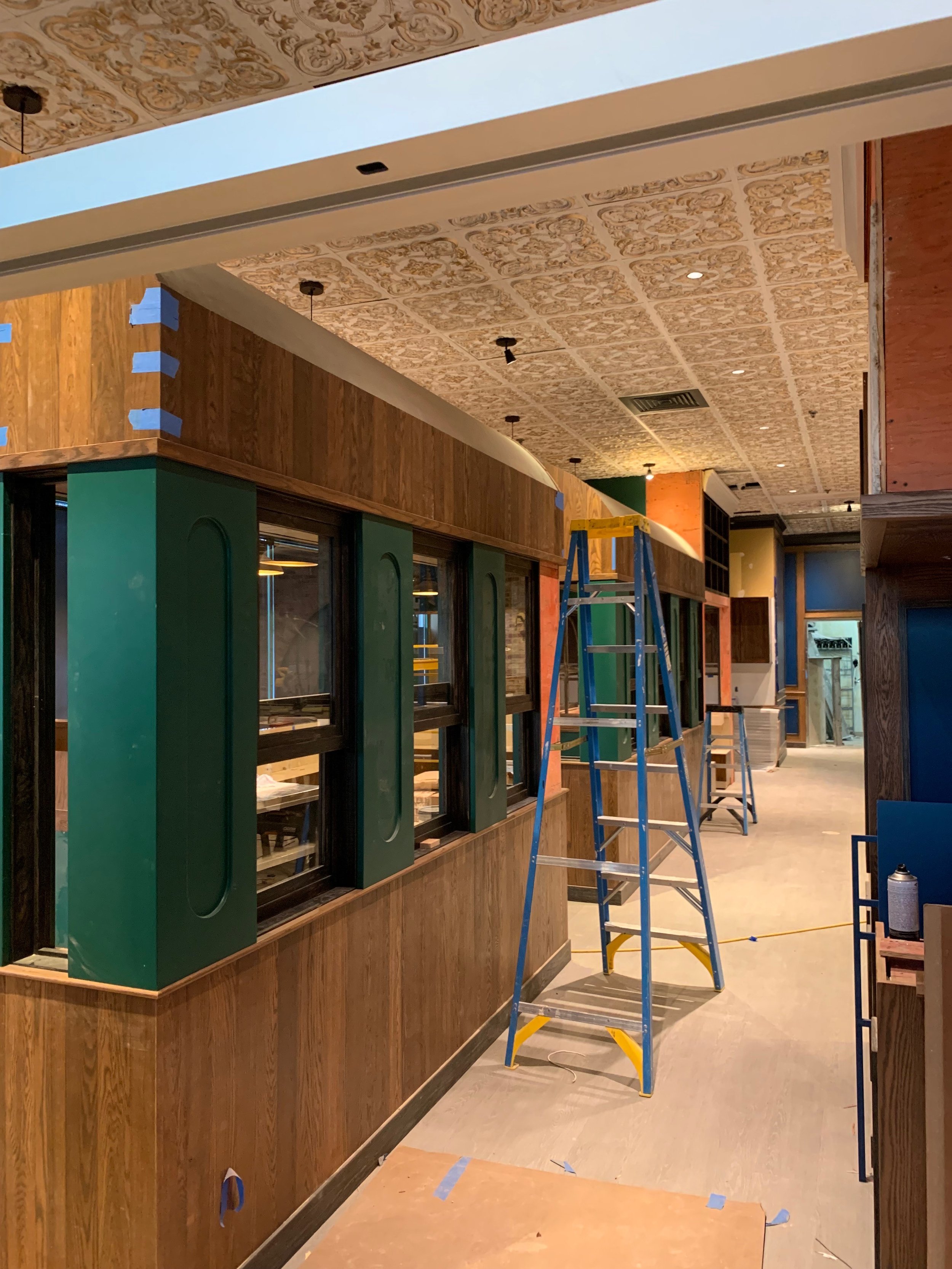 Interior of a restaurant or bar under renovation, with green and wood-paneled walls, dangling light fixtures, and a metal ladder in the middle of the space, with tape and construction materials scattered on the floor.