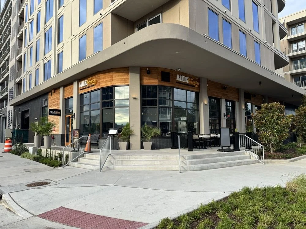 Exterior of a modern multi-story building with a restaurant called Milky Way, featuring large glass windows, outdoor seating, planters, and landscaping.