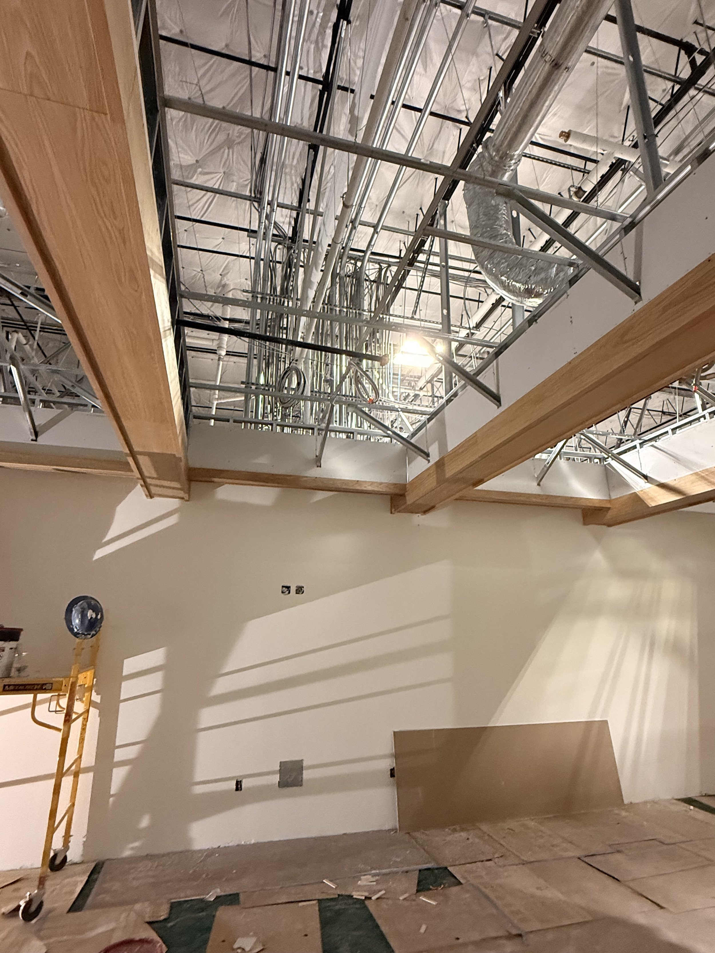 Inside of a building under construction showing exposed ceiling with pipes and ducts, wooden beams, and a partially finished wall with some wiring and construction tools.