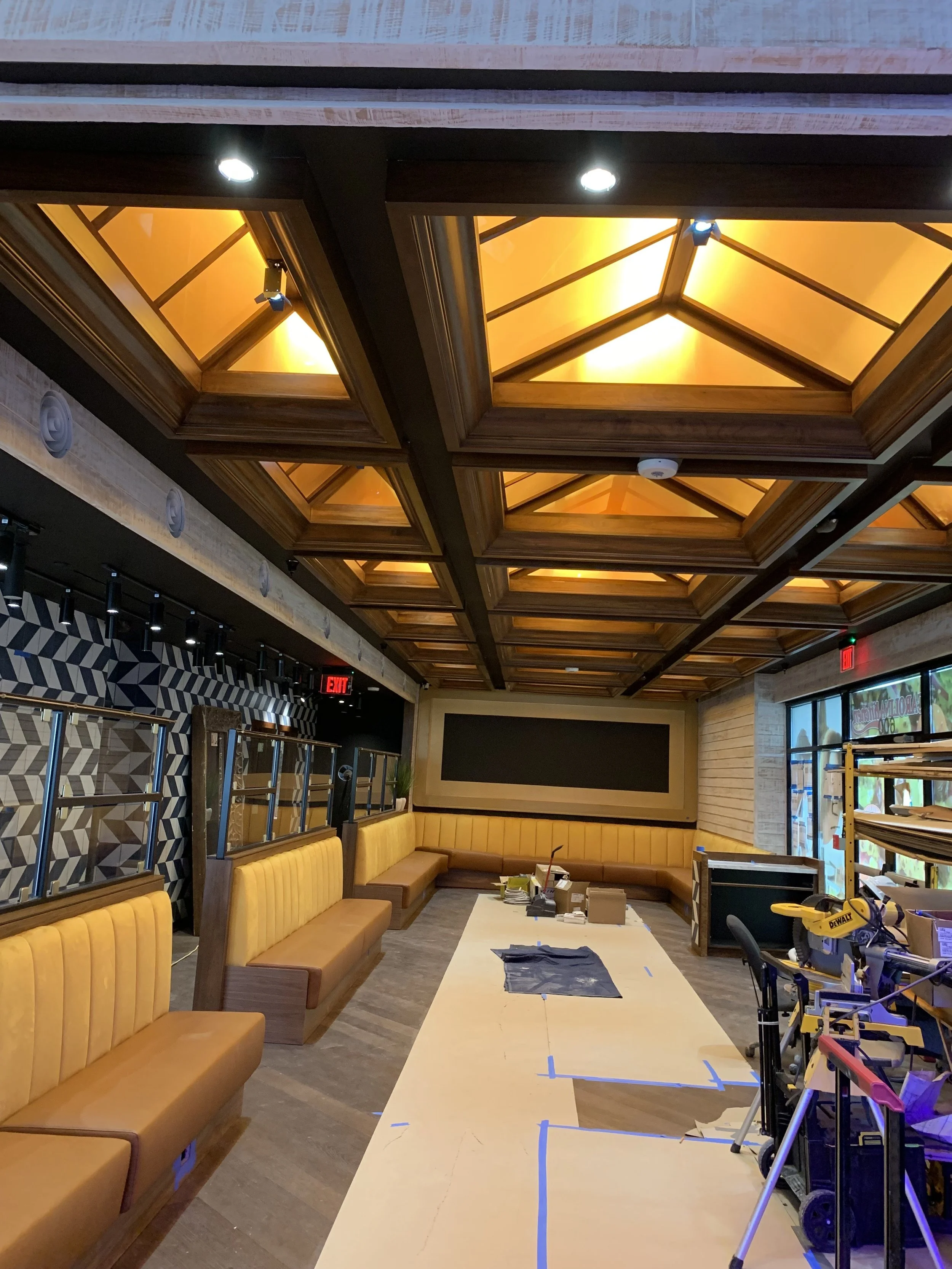 Interior of a restaurant or cafe under renovation with yellow booth seating along the walls, a long white work table in the center, and a ceiling with wooden beams and illuminated orange skylights. Construction tools and materials are visible.