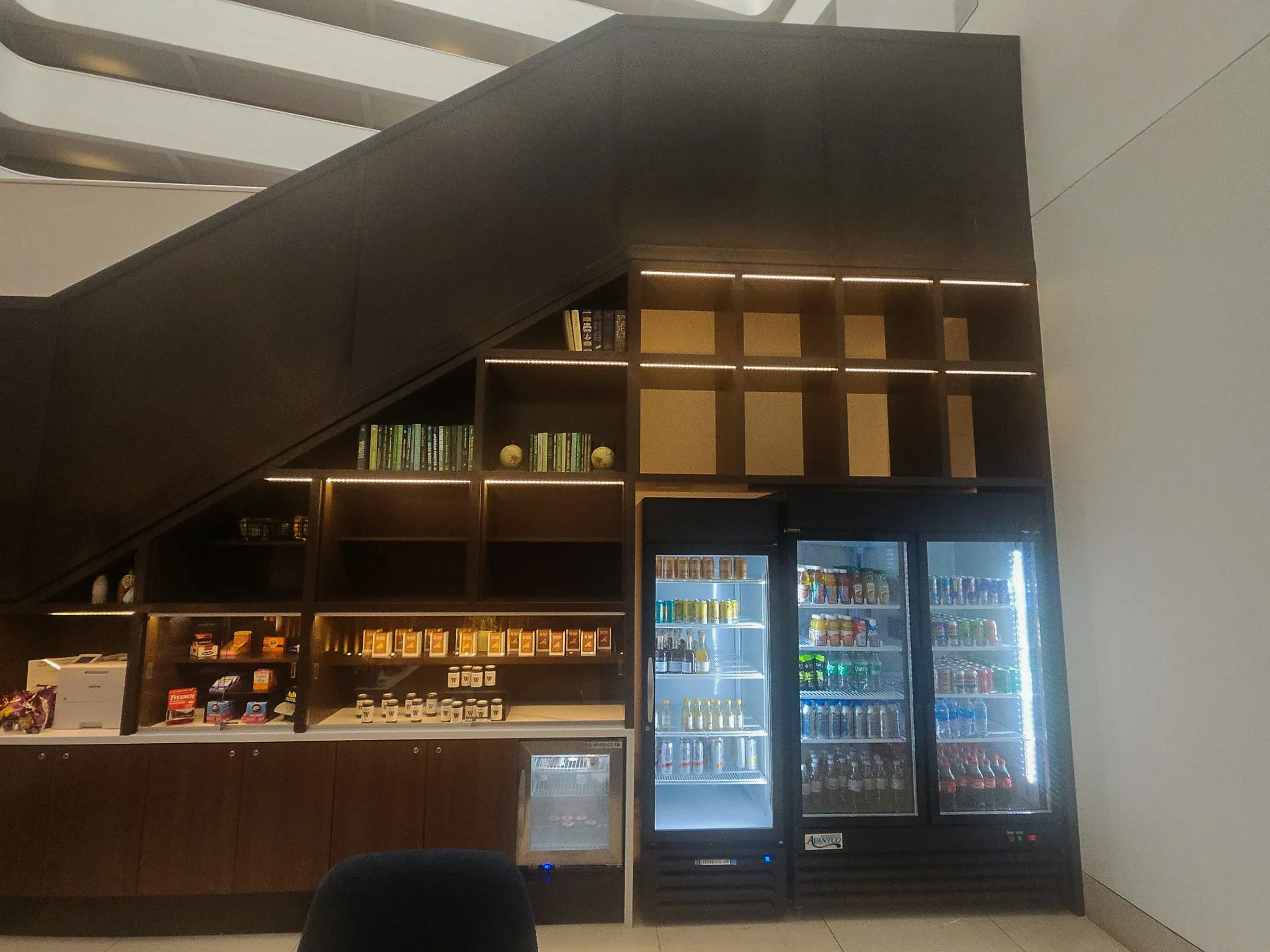 Image shows a refrigerated vending machine with beverages next to a wooden display shelf containing snack and beverage items, situated under a staircase with open risers in a modern interior.
