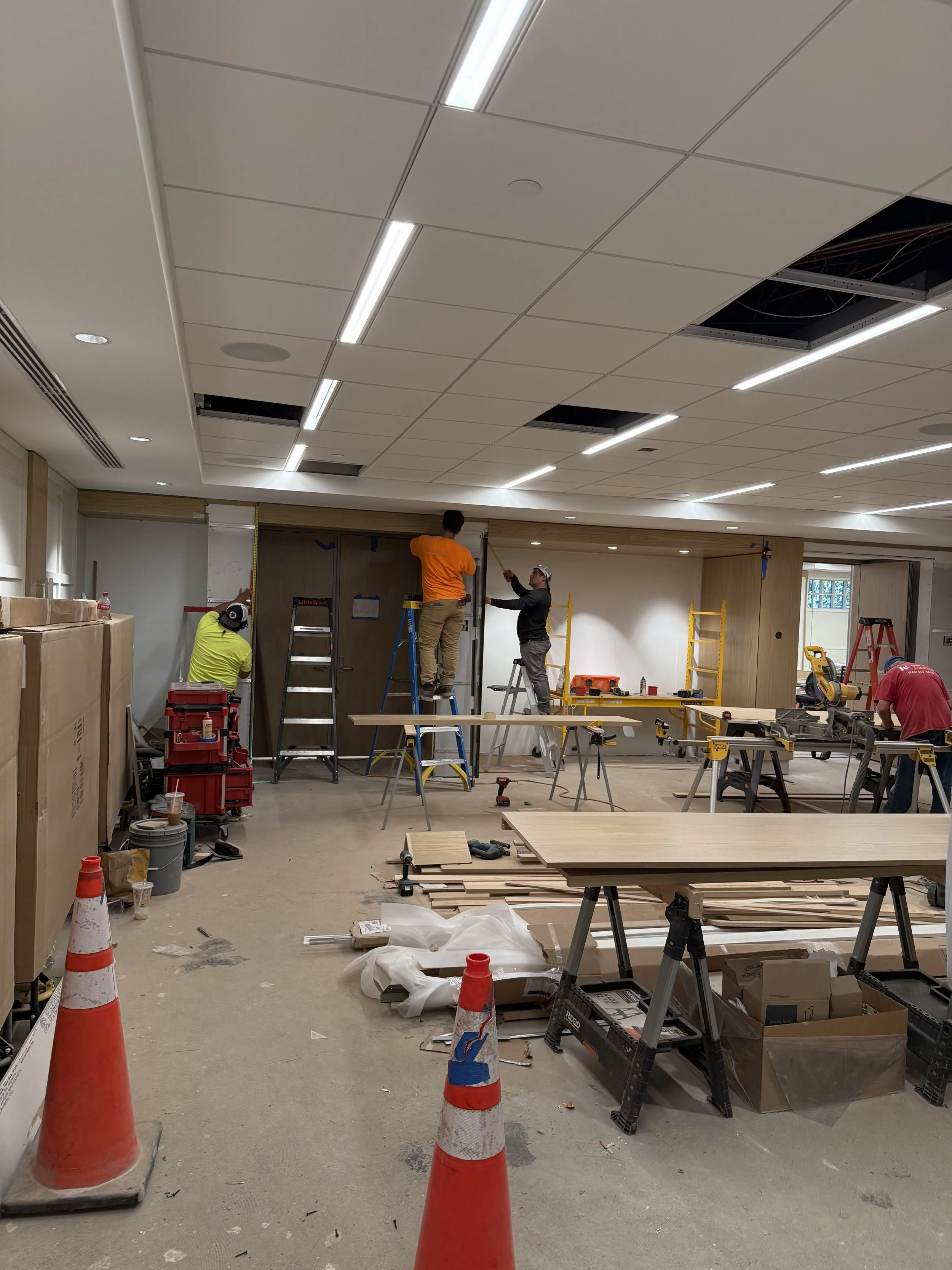 Construction workers installing ceiling panels in an indoor space, with tools, ladders, and materials scattered around.