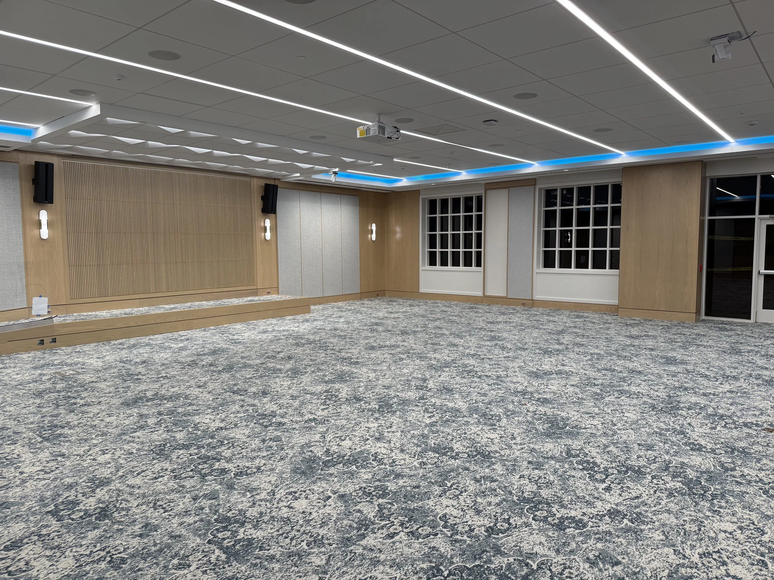 Empty modern conference or event room with carpeted floor, wood paneling on walls, large windows, and ceiling lighting.
