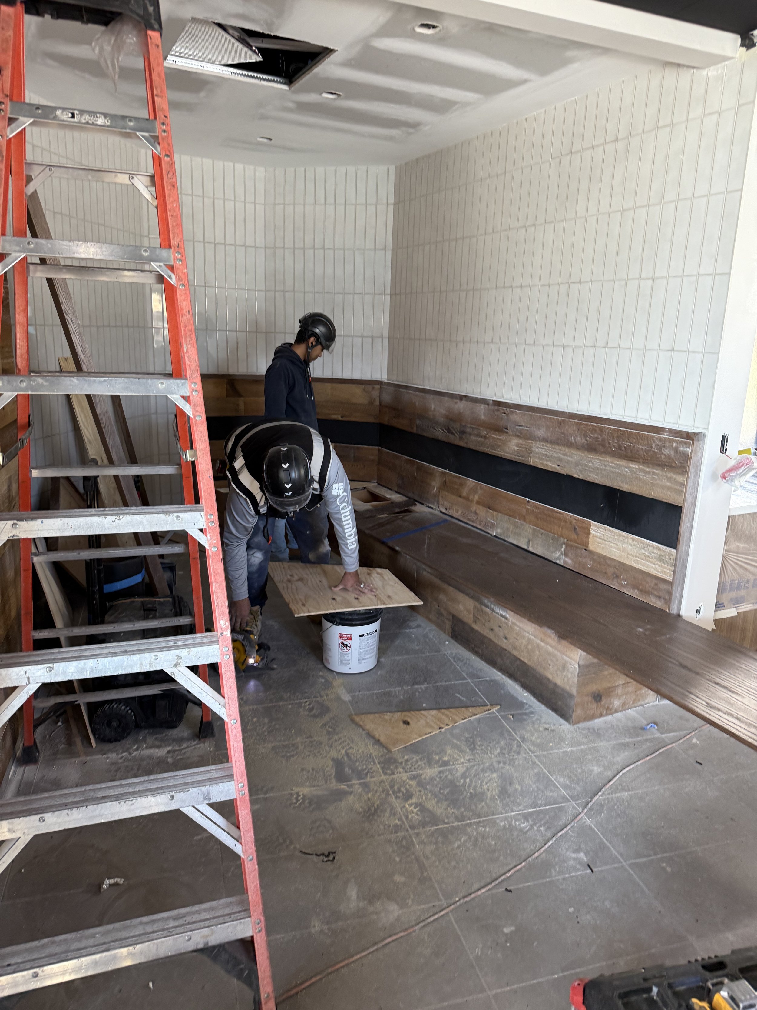 Construction workers installing wood paneling in an indoor space with tiled floor, a red ladder, and a partially finished speaker box.