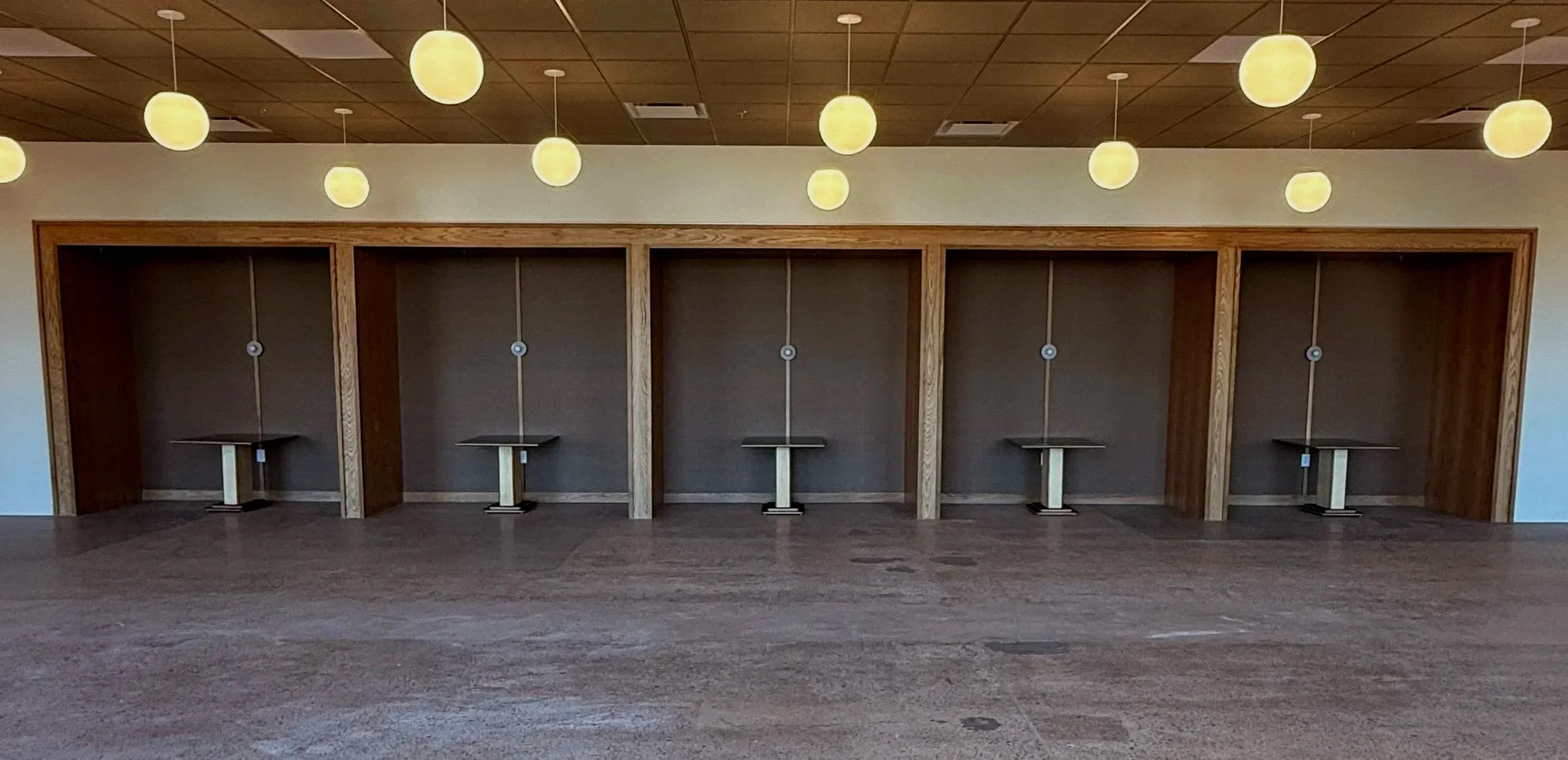 Six empty privacy booths with small tables, separated by wooden frames, positioned along a wall in a room. Overhead lighting fixtures with round, yellowish shades hang from the ceiling.