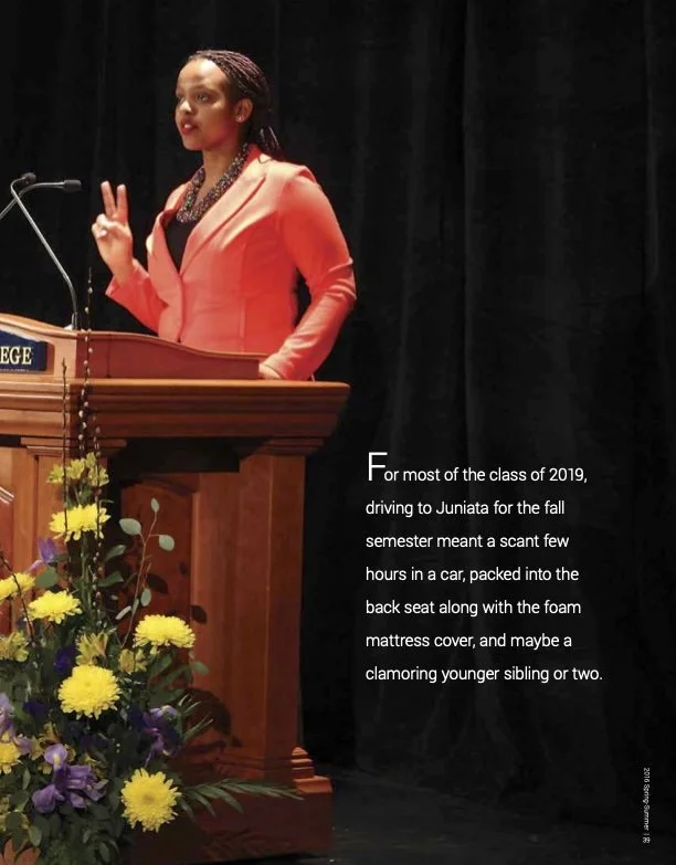 A woman wearing a coral blazer stands behind a wooden podium, making a peace sign with her right hand during a speech or presentation. Feature article news writing reporting creative writing.