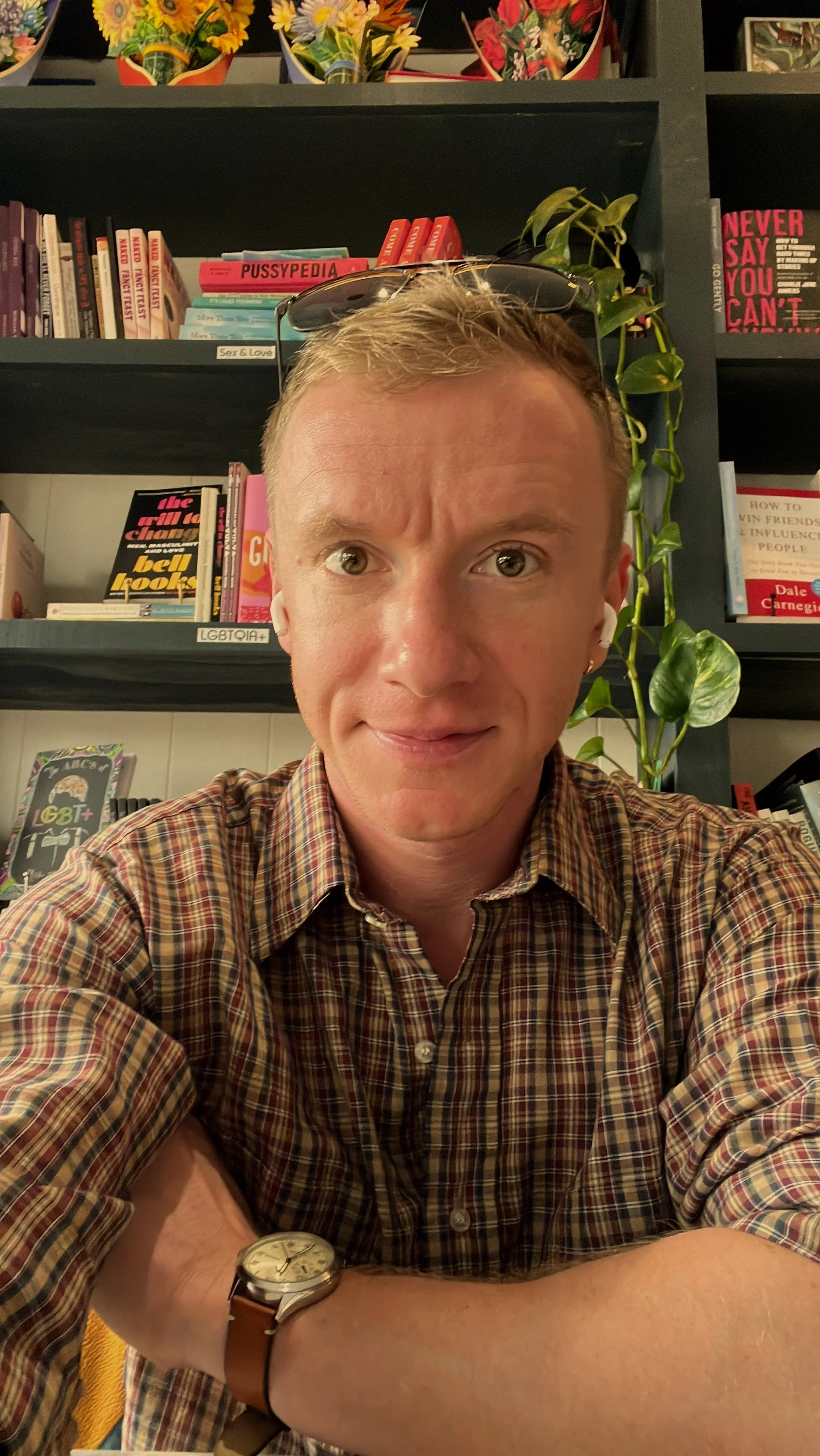 A man with short blond hair wearing a plaid shirt and a watch, sitting in front of bookshelves filled with various books, plants, and decorative items. Fiction creative writing Tyler Ayres.