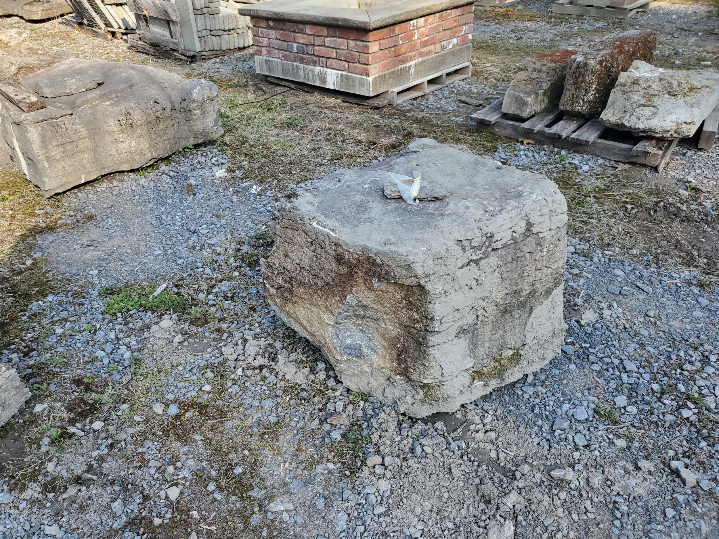A large square stone block with a small rock and a partially torn piece of paper on top, in an outdoor yard with gravel, rocks, and construction materials