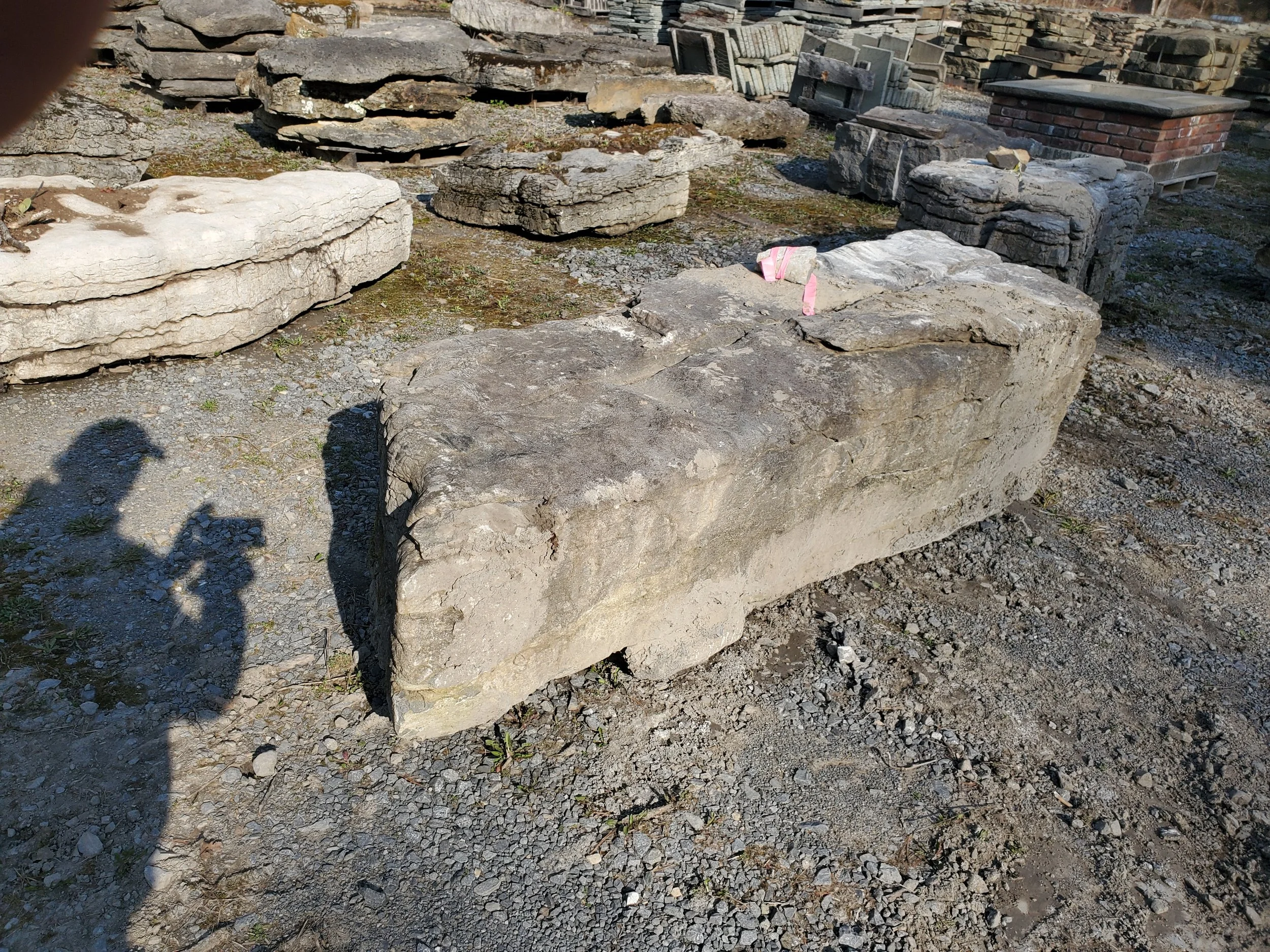 Large, irregularly shaped stone block on gravel ground in a stone yard with various stacked stones and bricks in the background.
