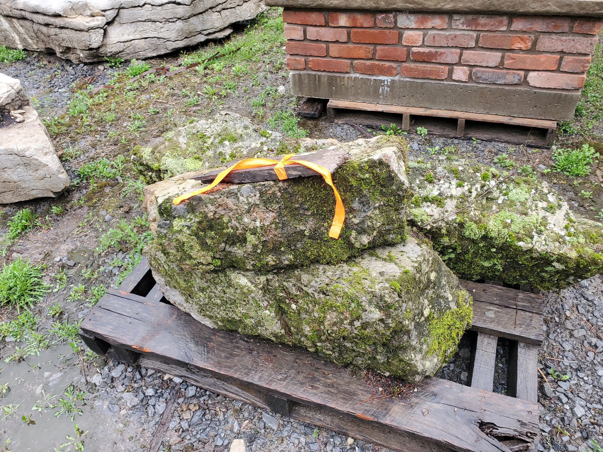A pile of large moss-covered rocks stacked on a wooden pallet in an outdoor area with dirt, small green plants, and a brick building in the background.