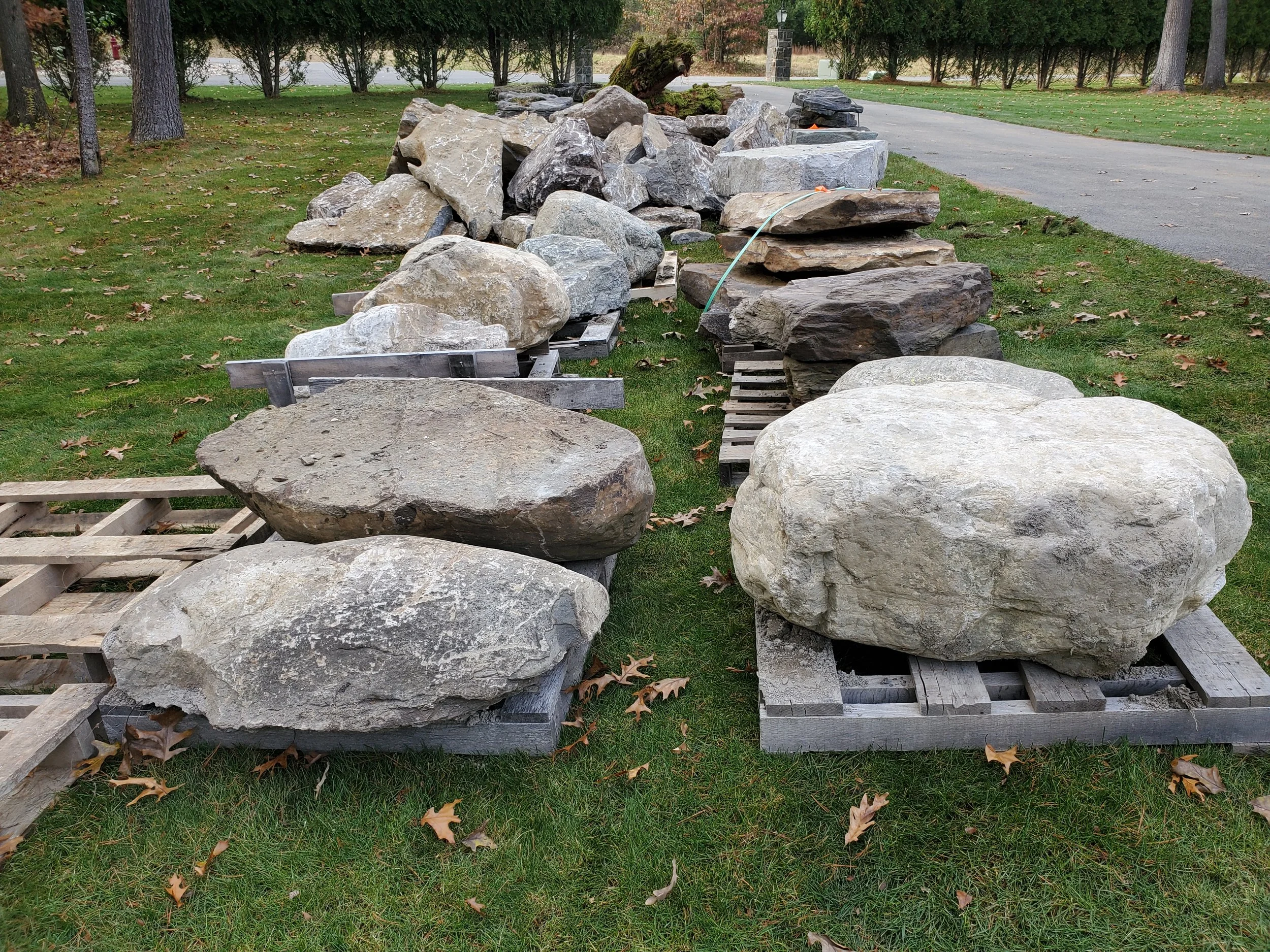 Large rocks arranged on wooden pallets outdoors on a grassy area, with trees and a road in the background.