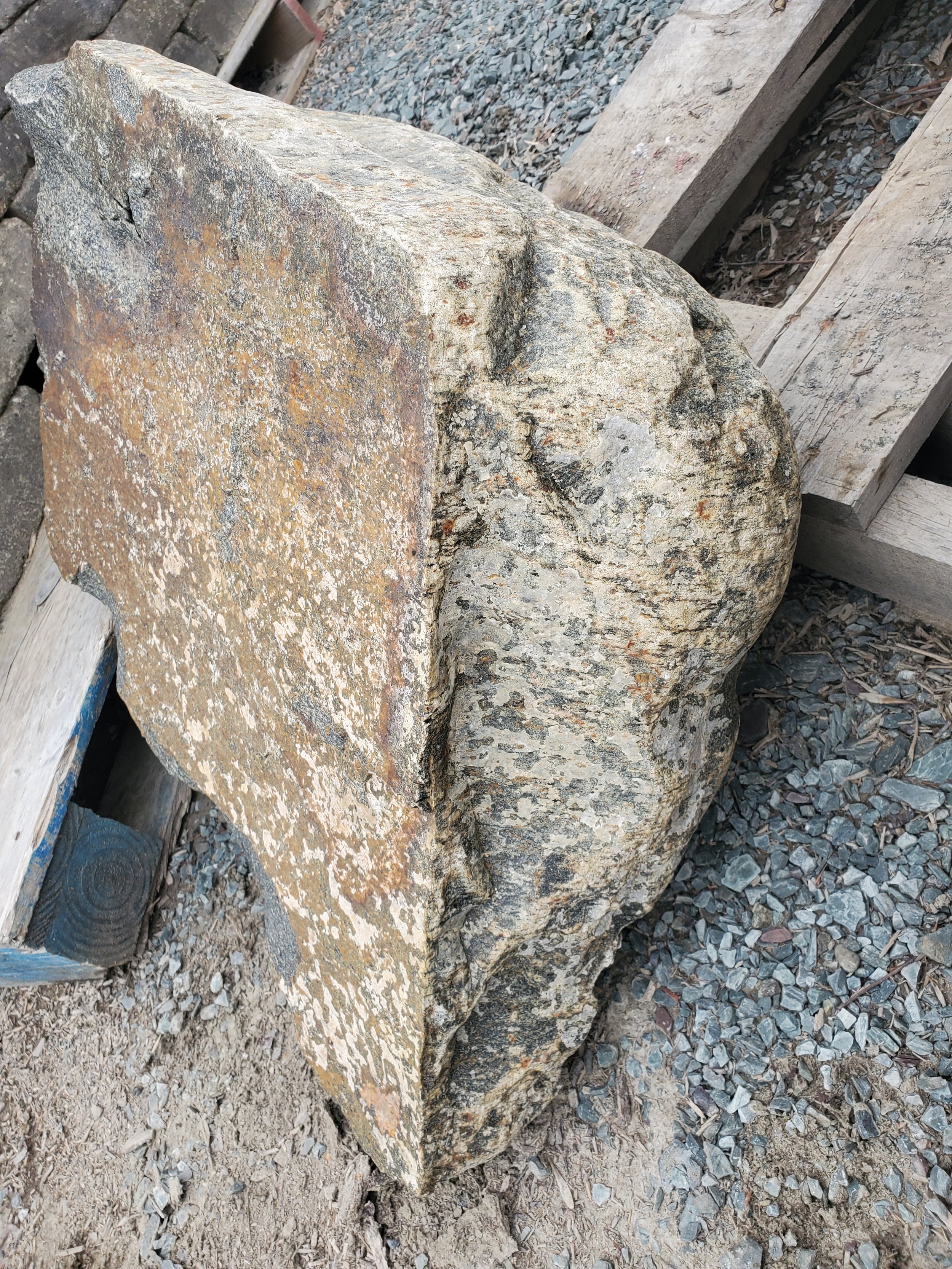 A large, irregularly shaped rock with a rough, textured surface resting on gravel and dirt, surrounded by wooden planks.