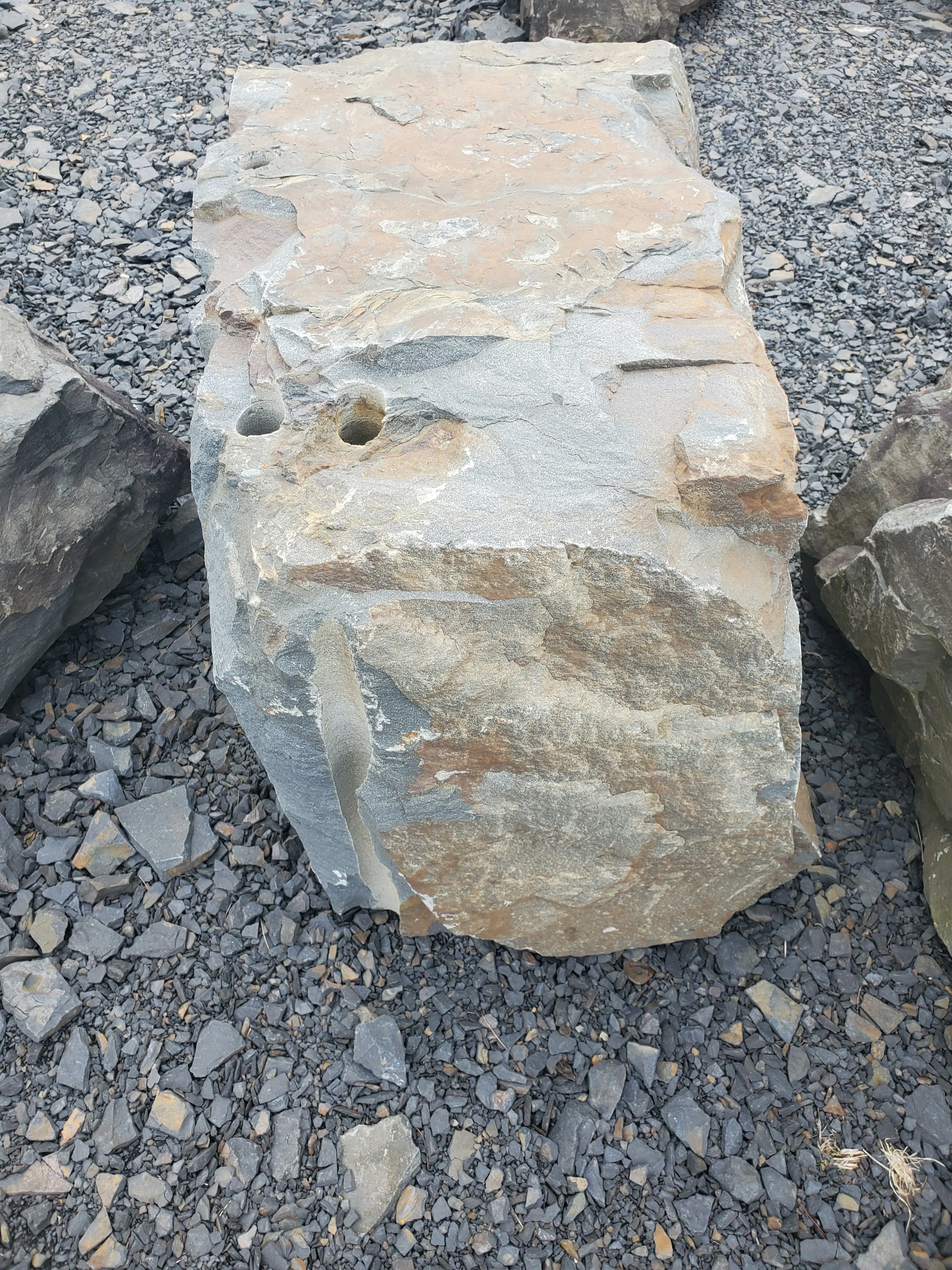 Large irregularly shaped stone with small holes on its top surface, placed on gravel.