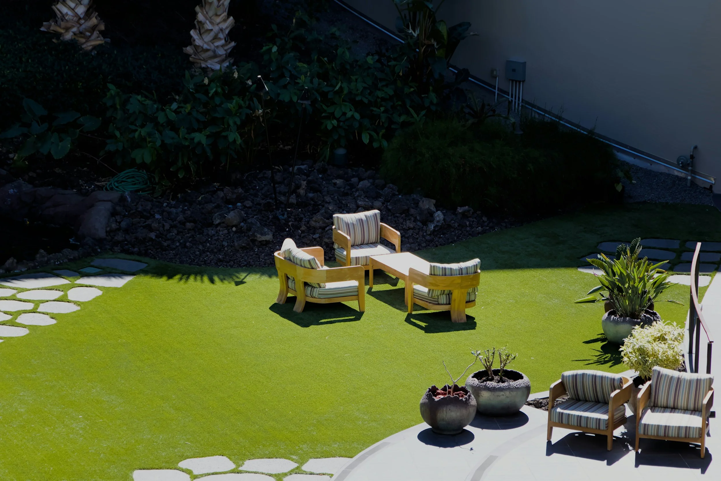 A backyard with artificial grass, outdoor chairs with striped cushions, potted plants, and stone pathway